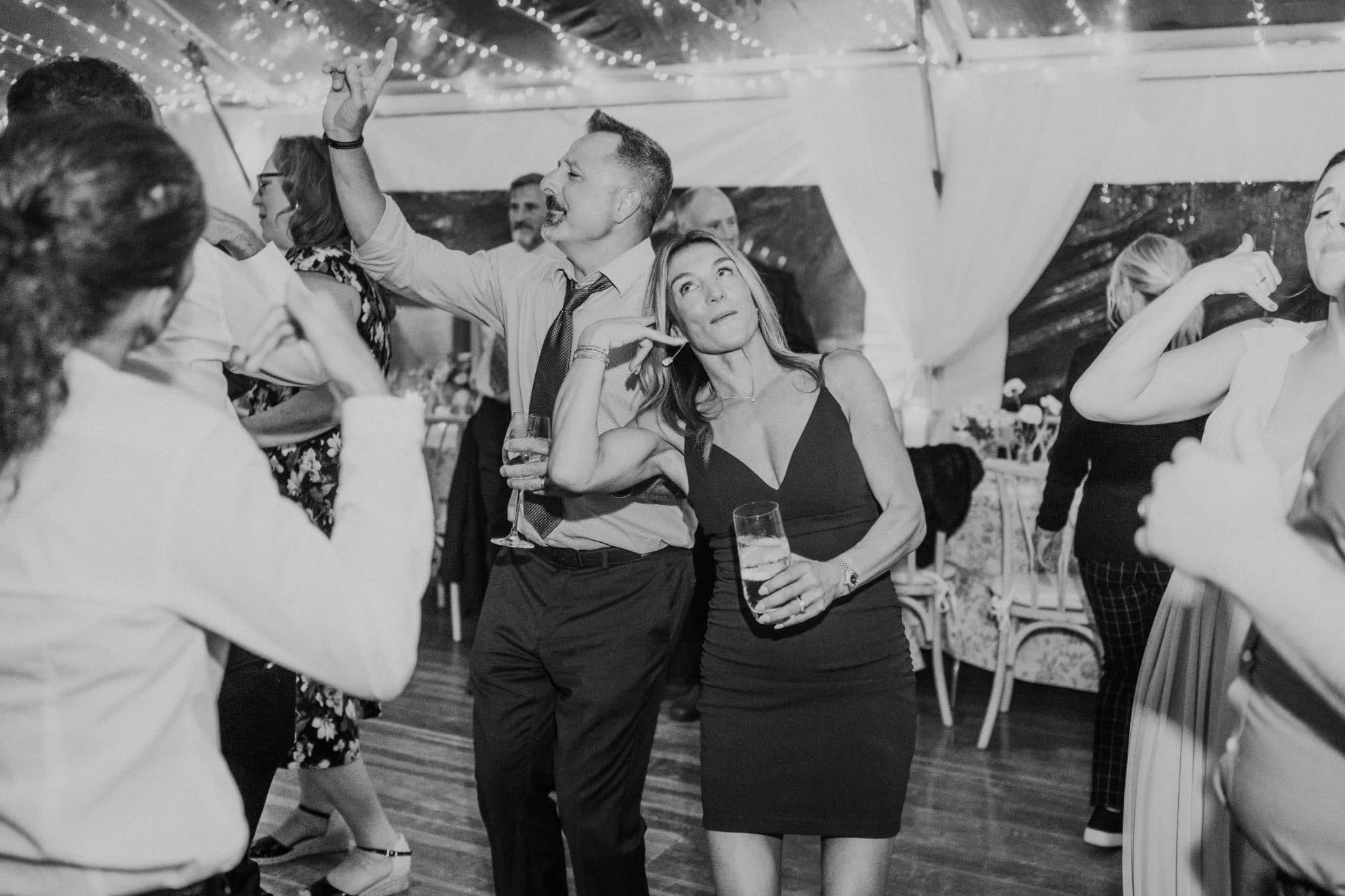 A black and white wedding photograph captures a lively dance floor with guests in formal attire, holding drinks, and enjoying the celebration.