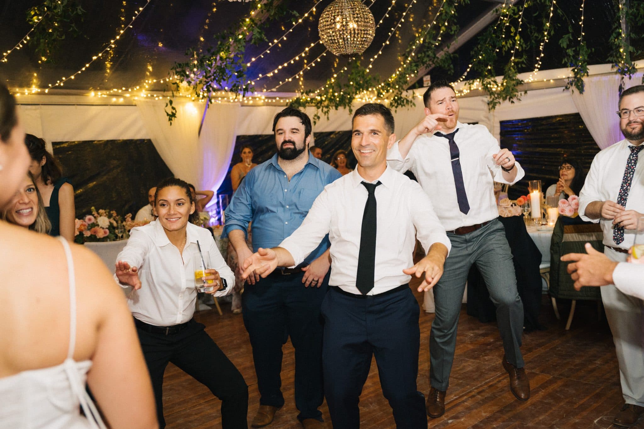 A group of people in formal attire dances joyfully under string lights and a chandelier at a wedding reception.