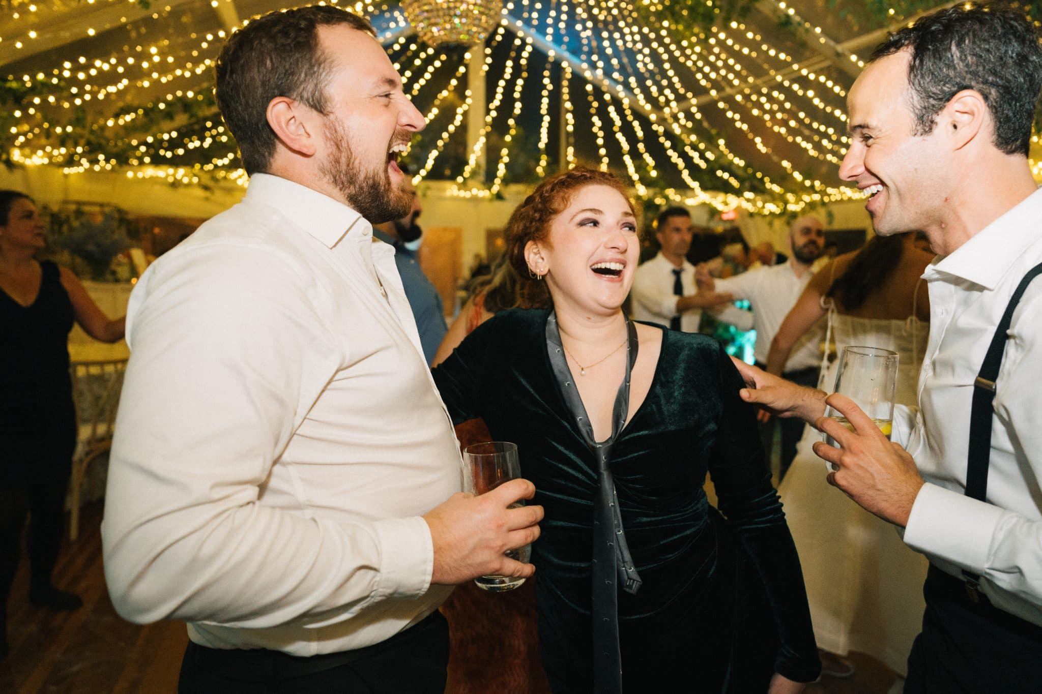 Three people laugh together at a festive event with string lights in the background.