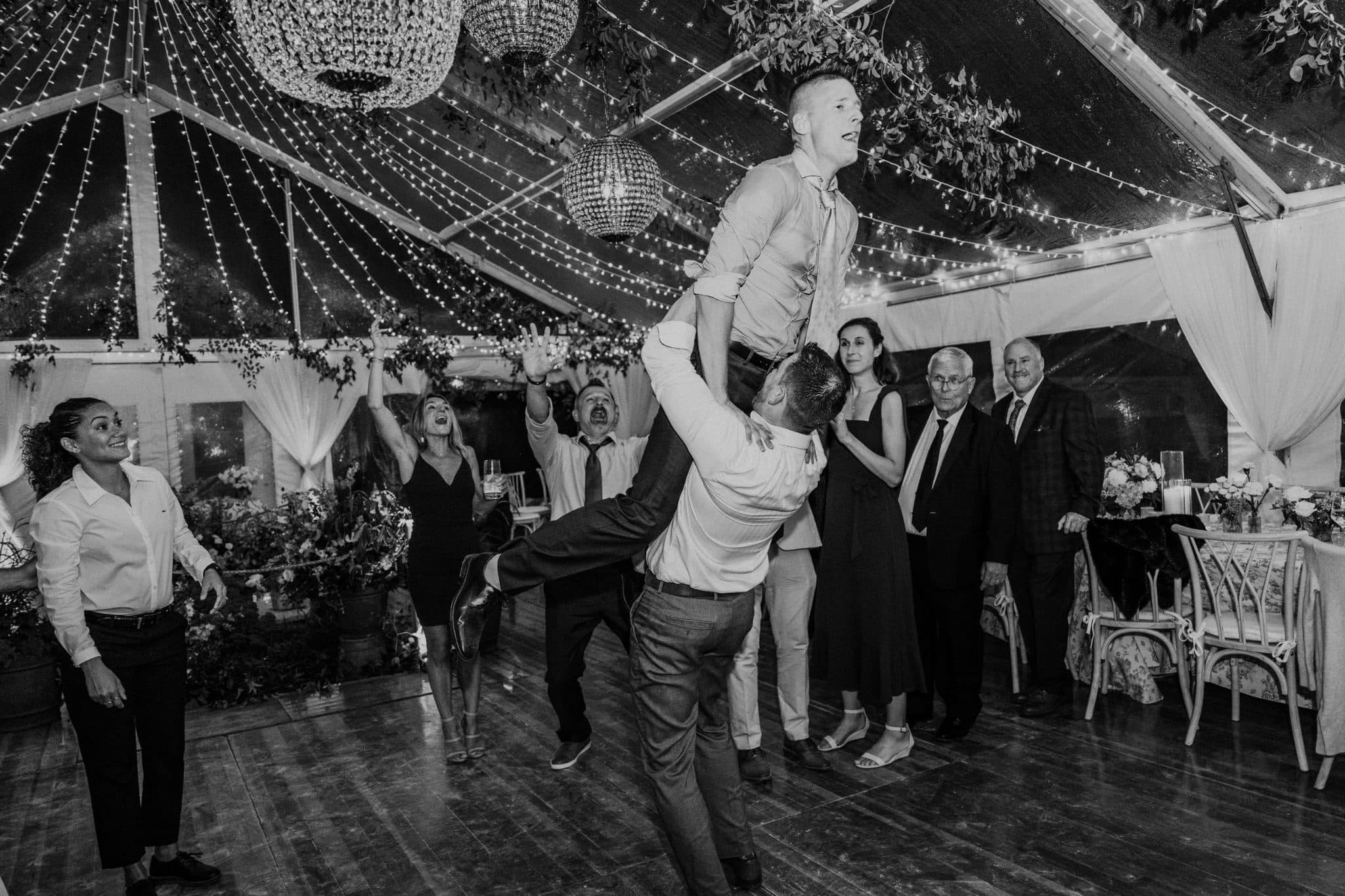A man is being lifted in the air by another man at a wedding reception under a tent decorated with lights and flowers.