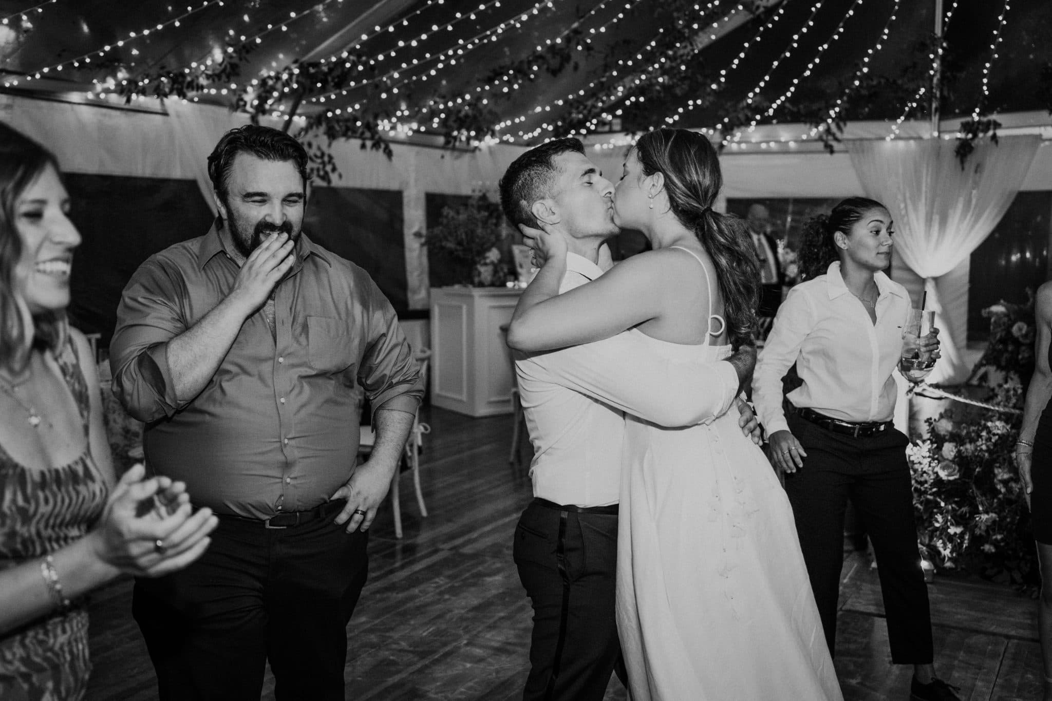A couple kisses on a dance floor surrounded by guests at a wedding reception.