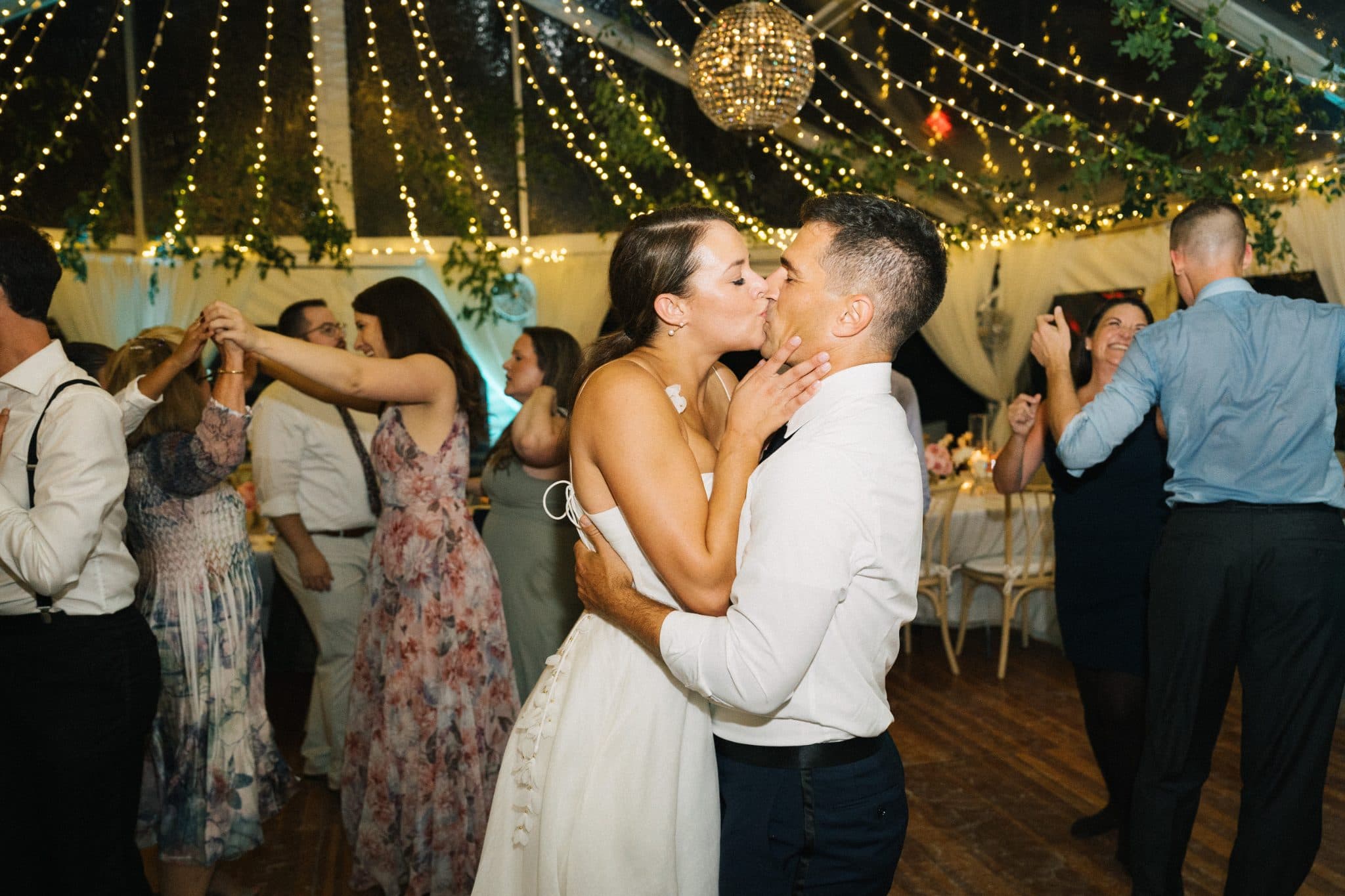 A couple in wedding attire shares a kiss on the dance floor amidst guests and string lights.