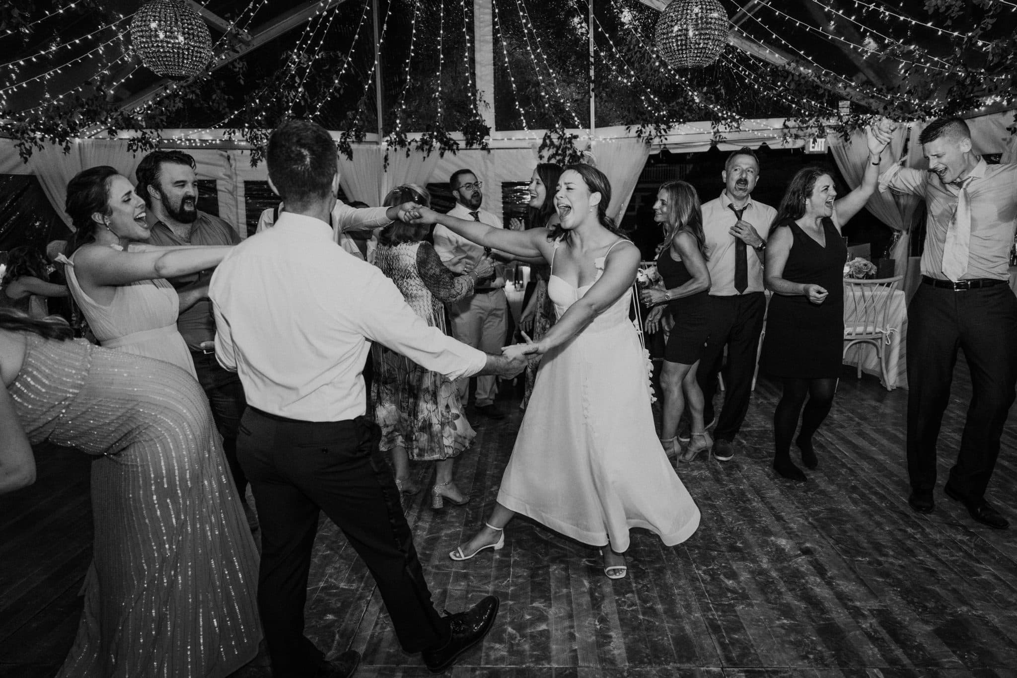 A bride and groom dance joyfully under a tent adorned with lights and greenery.