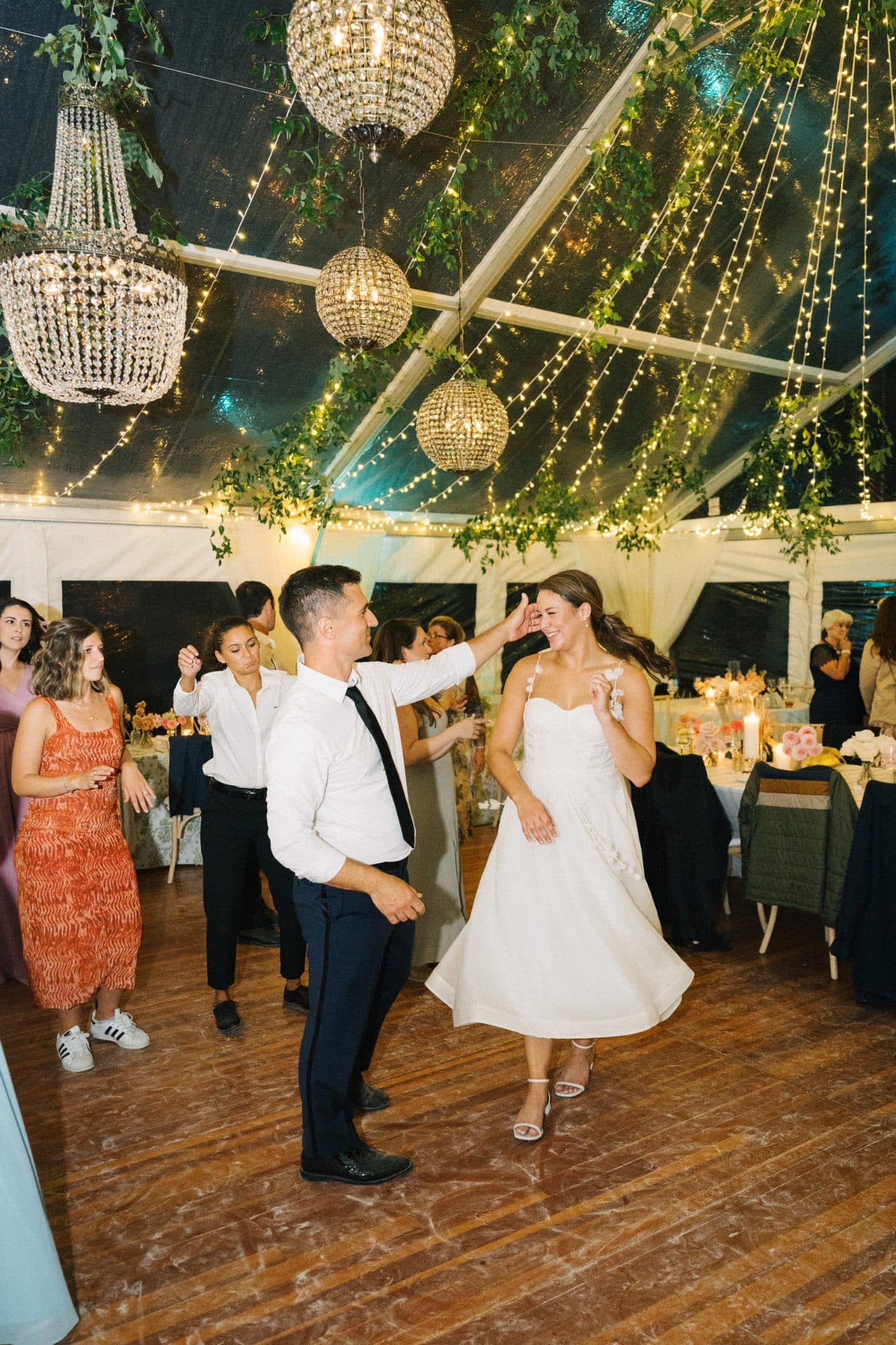 A bride in a white dress dances with a man in a white shirt and black tie under a tent adorned with hanging lights and greenery.