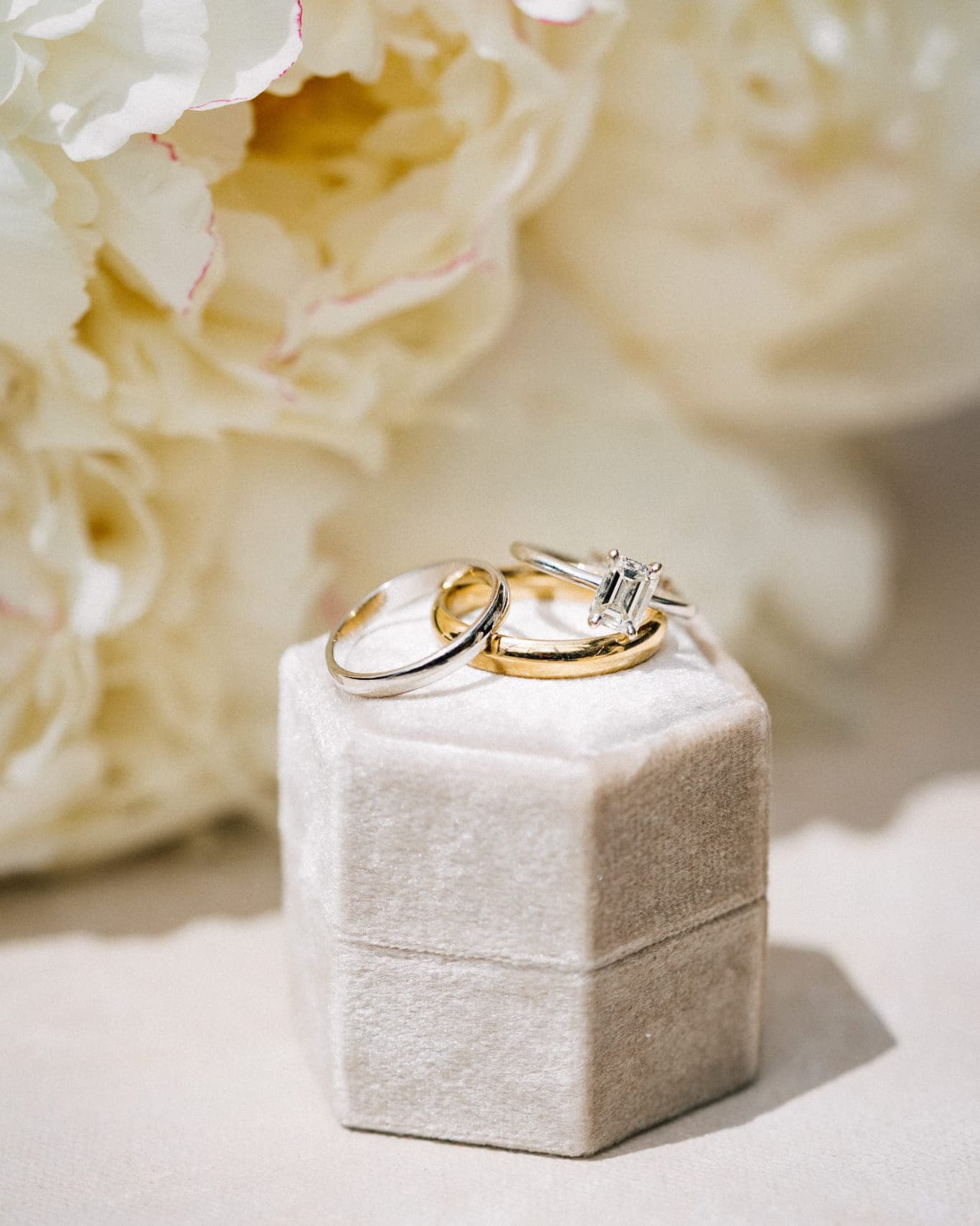 A wedding ring and engagement ring sit atop a small, white, velvet box with a blurred floral background.
