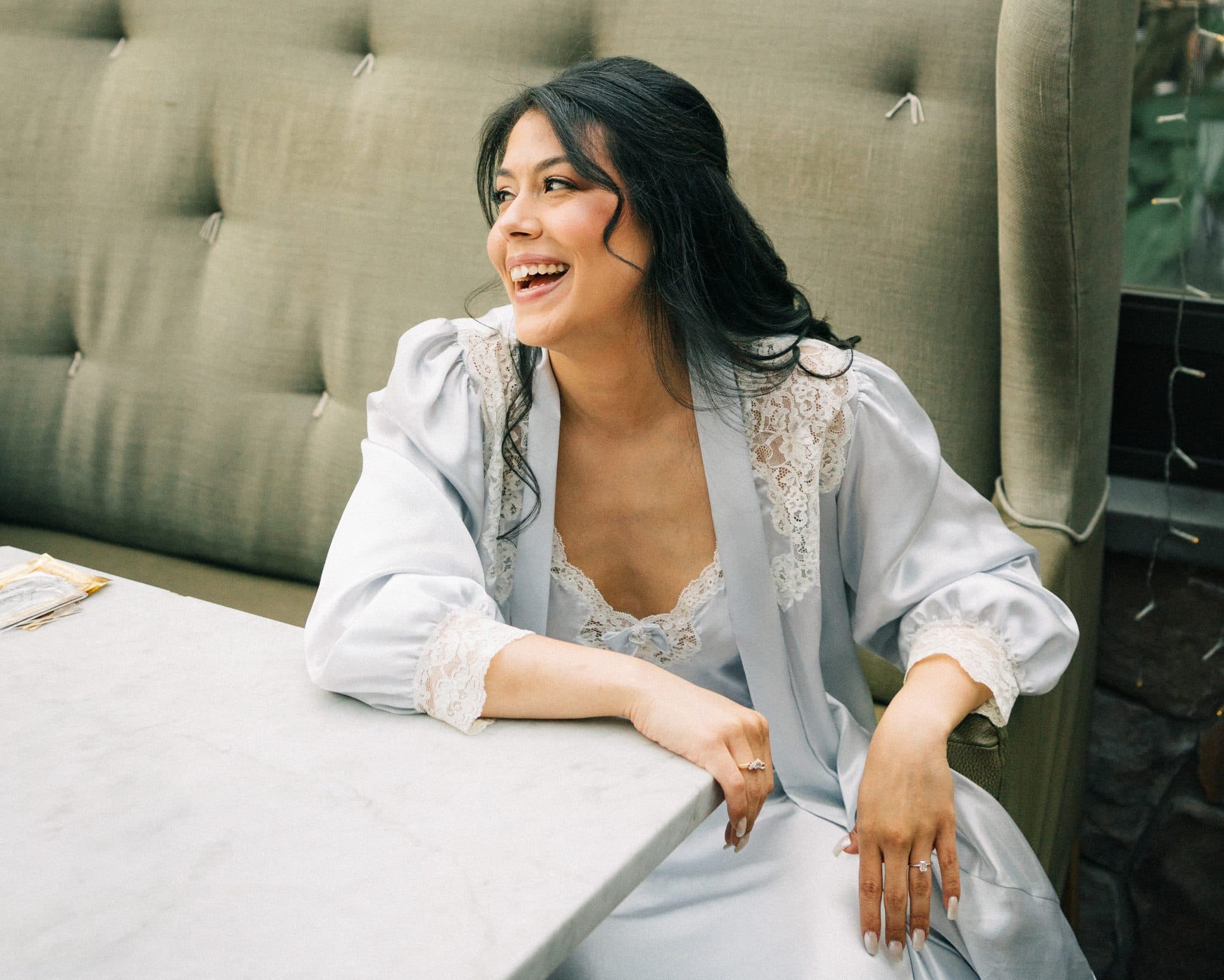 A woman in a light blue robe with lace details sits at a table, smiling with her hands resting on the table.