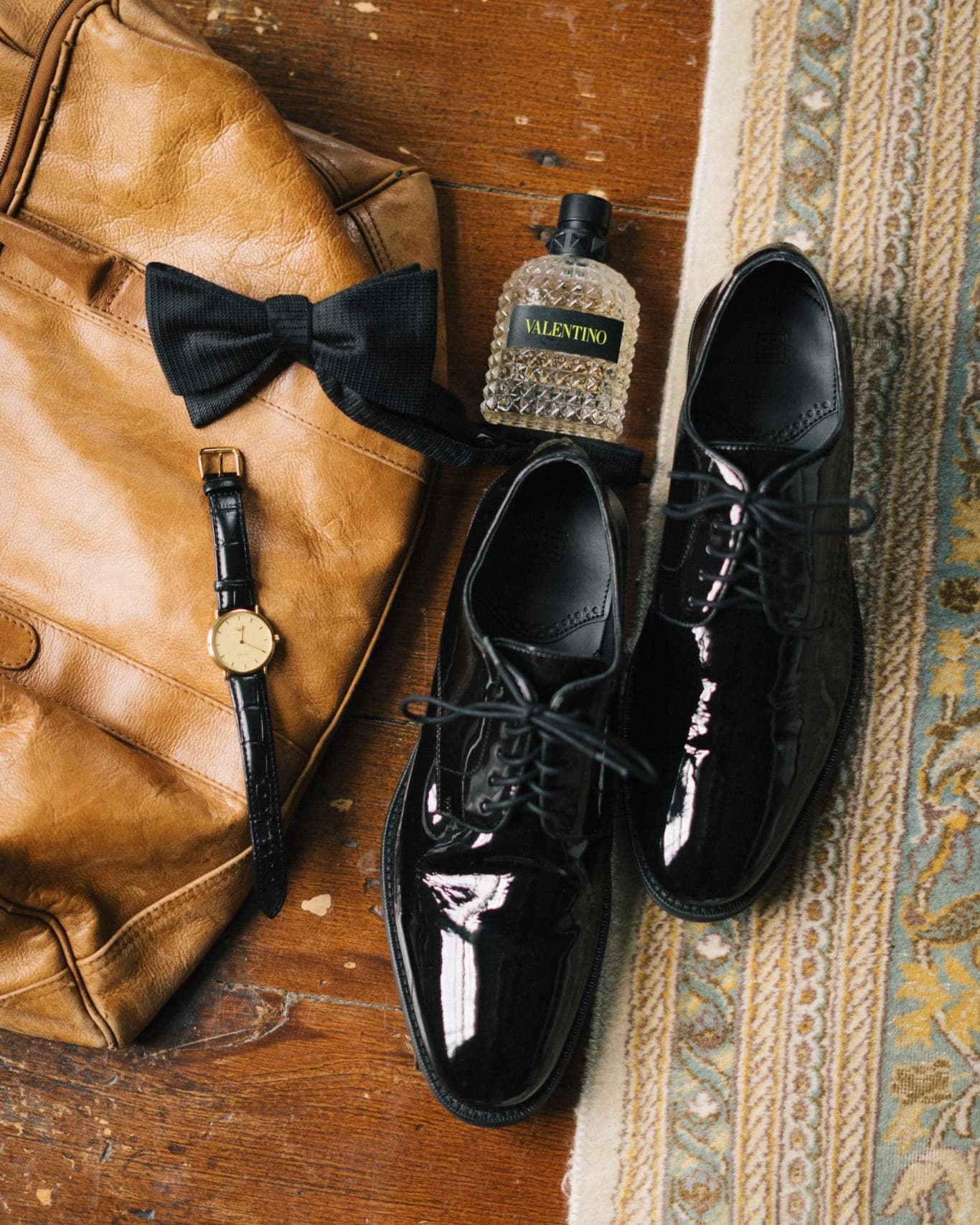 A brown leather jacket, black bow tie, watch, black dress shoes, and a perfume bottle are arranged on a wooden floor with a patterned rug.