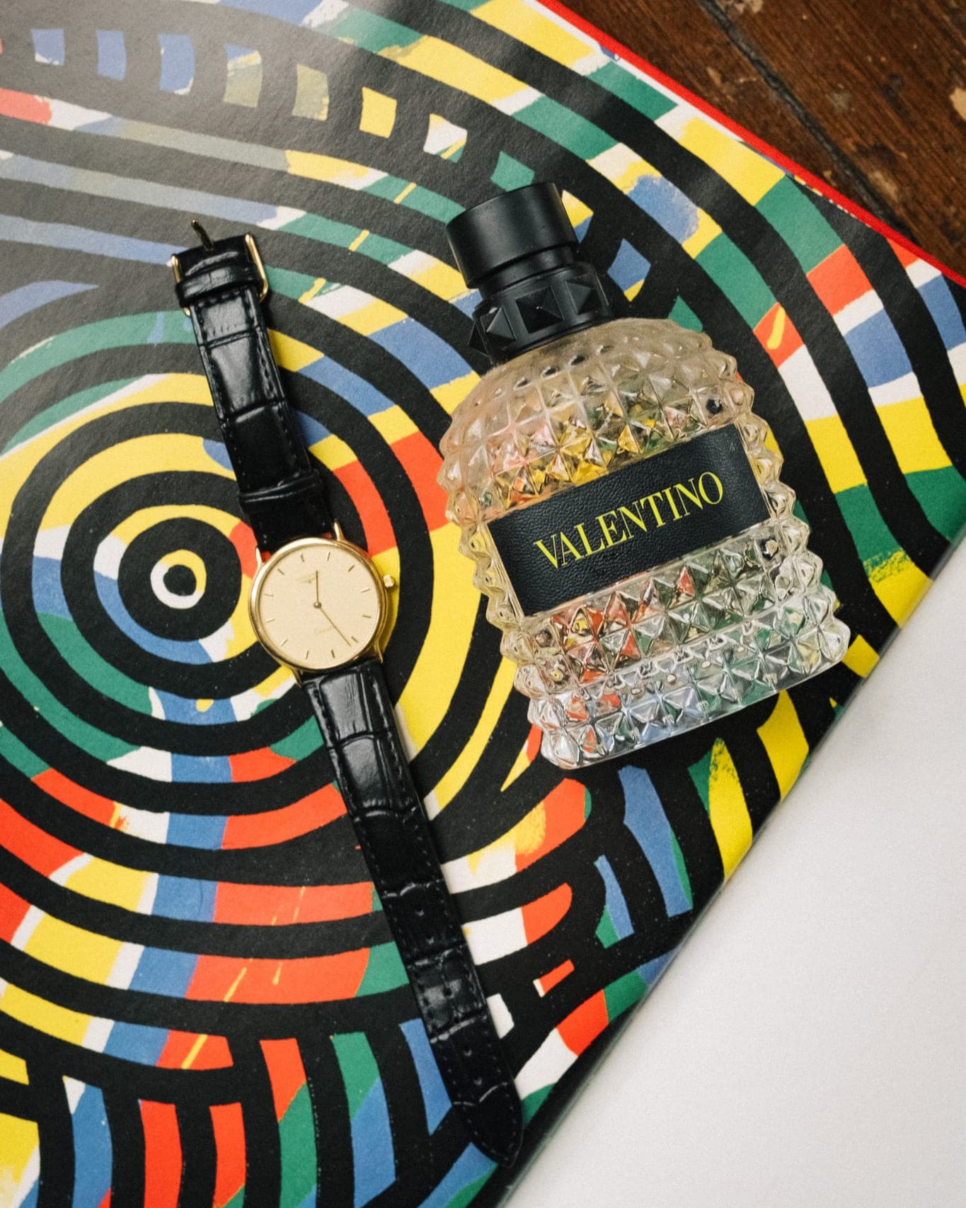 A watch with a black leather strap and a perfume bottle with a black cap and a textured glass body sit on a colorful, abstract patterned surface.