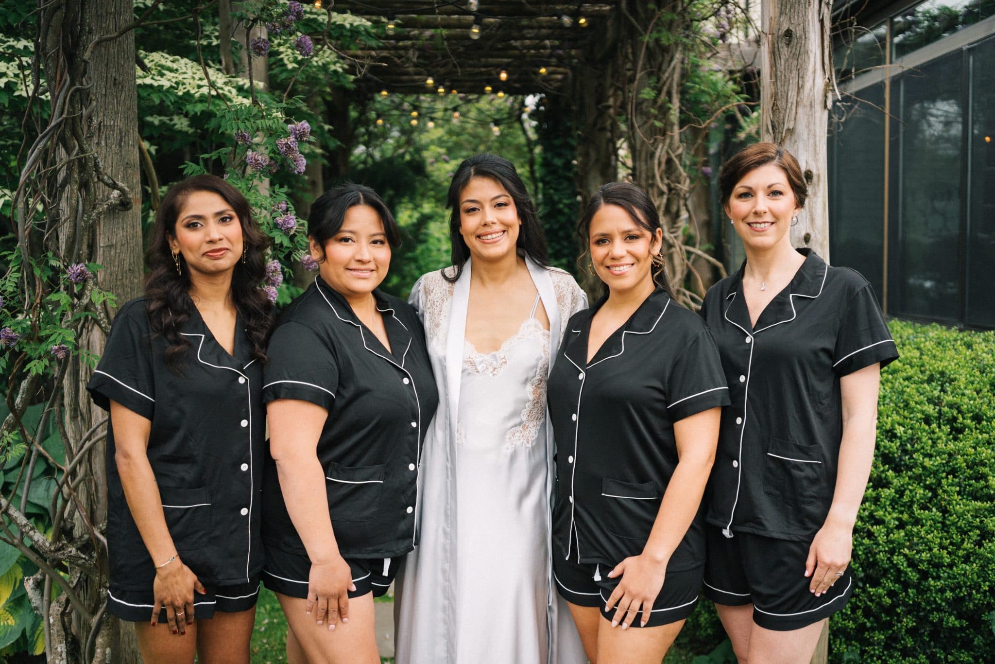 Five women in matching black pajamas stand together, one in a white dress, in a garden setting with greenery and hanging lights.