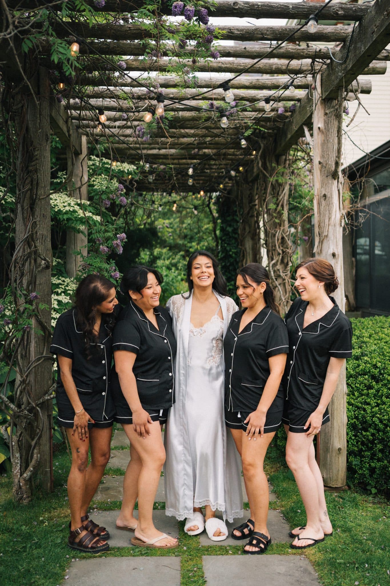A bride in a white robe stands with four women in black pajamas under a wooden pergola with hanging lights and purple flowers.