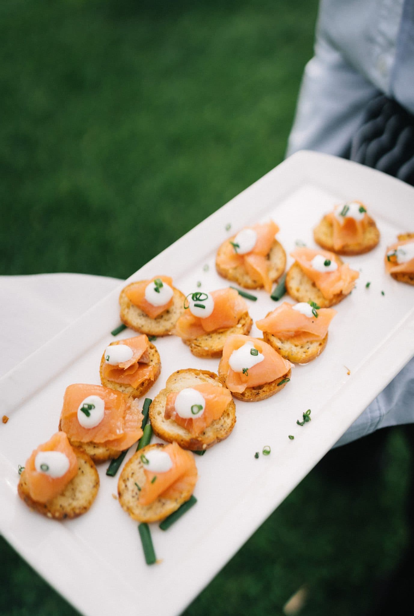 A white rectangular plate holds small crostini topped with smoked salmon, cream cheese, and chives.