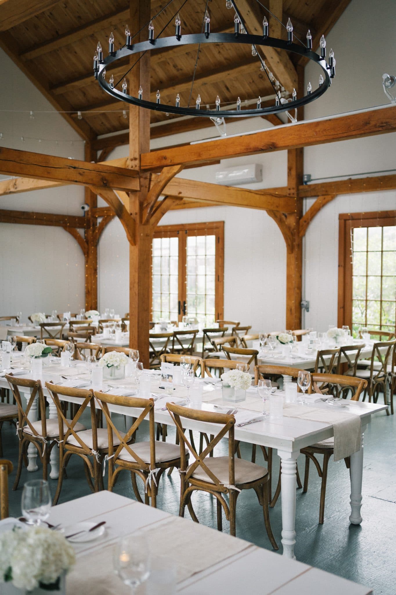 A warmly lit wedding reception with white tables, wooden chairs, and floral centerpieces in a rustic barn setting.