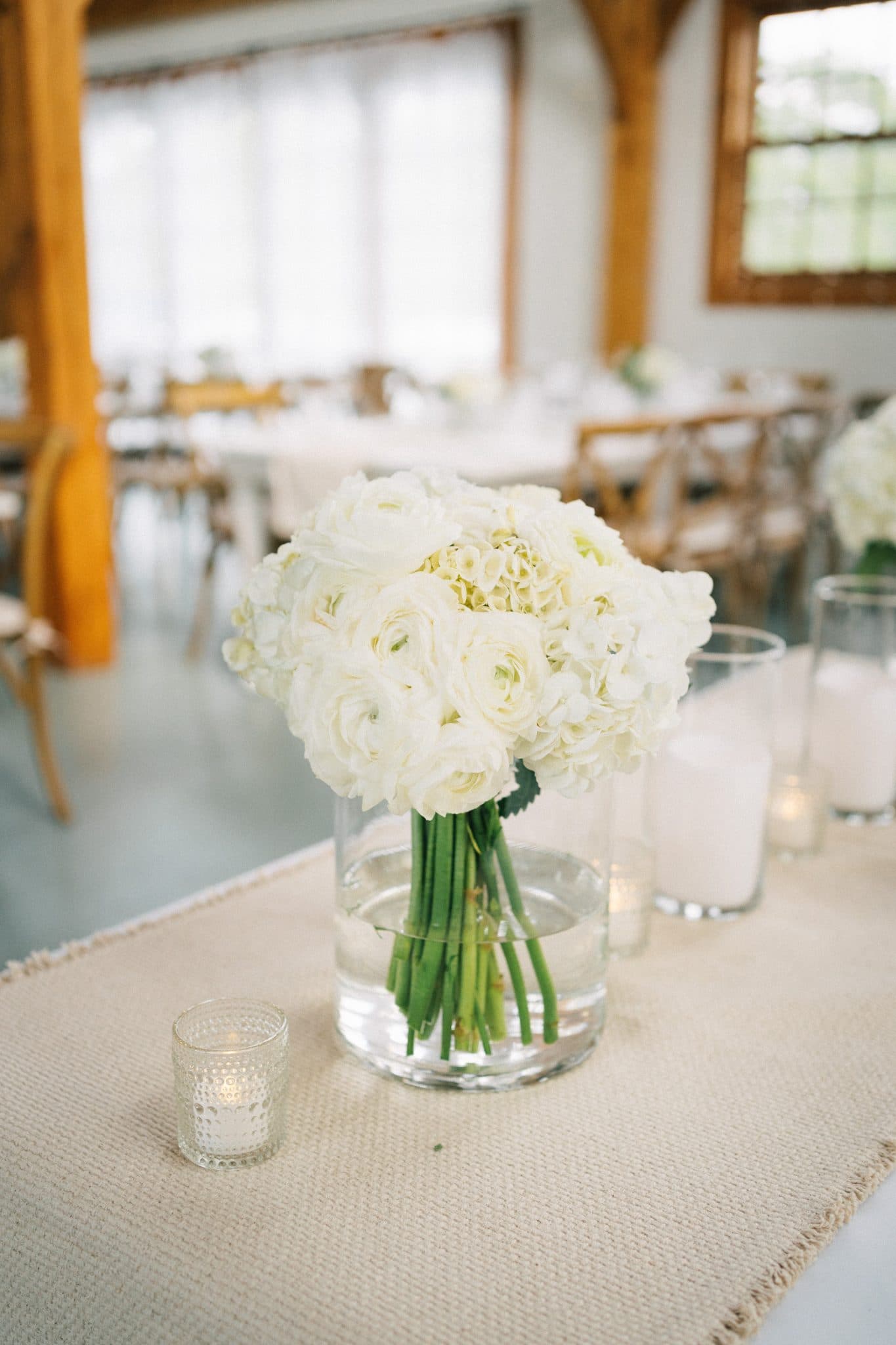 A table setting features a bouquet of white flowers in a glass vase, surrounded by lit candles and a textured tablecloth.