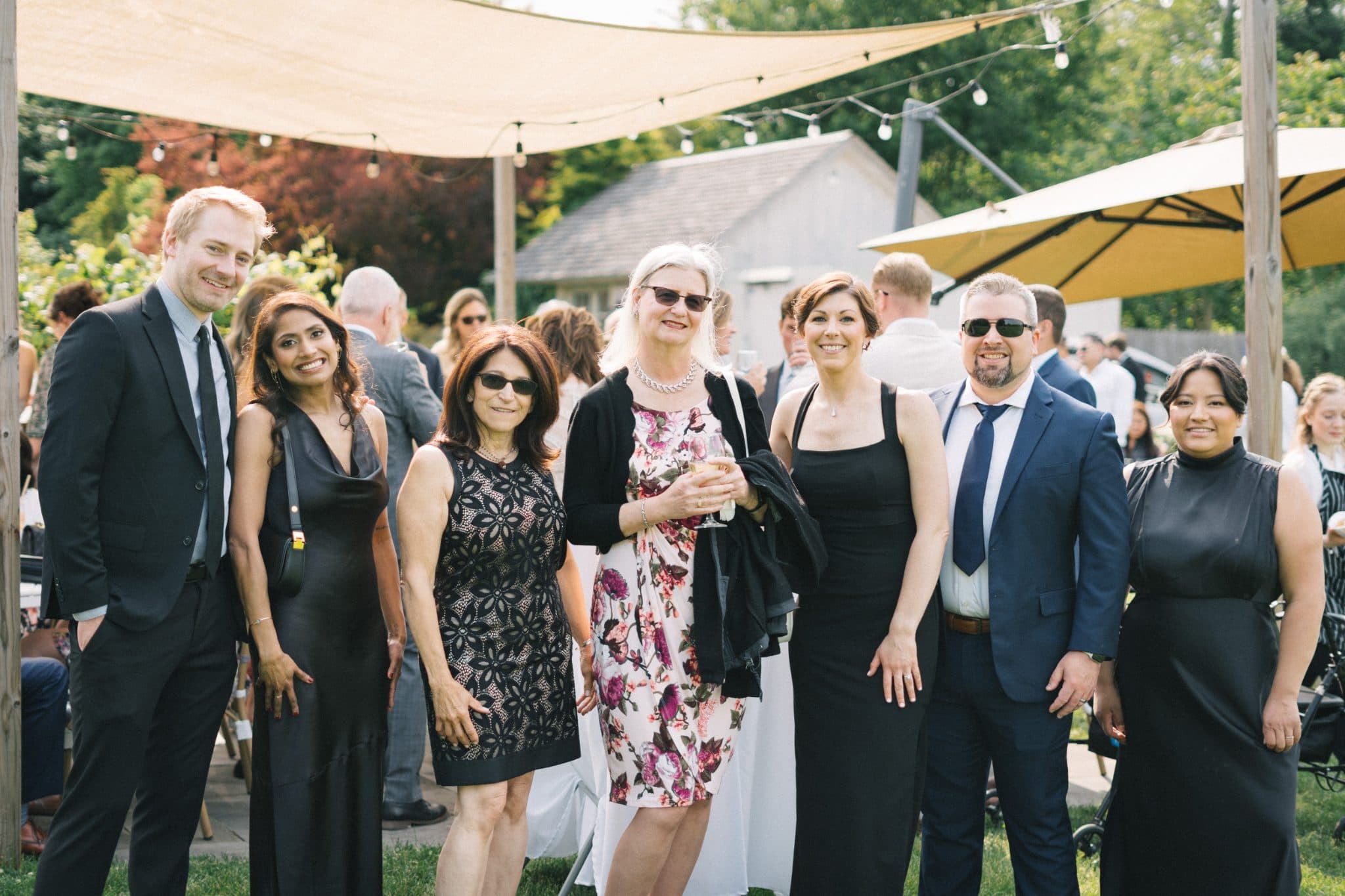 A group of seven people, dressed in formal attire, stand together outdoors under a canopy, smiling and posing for a photograph.