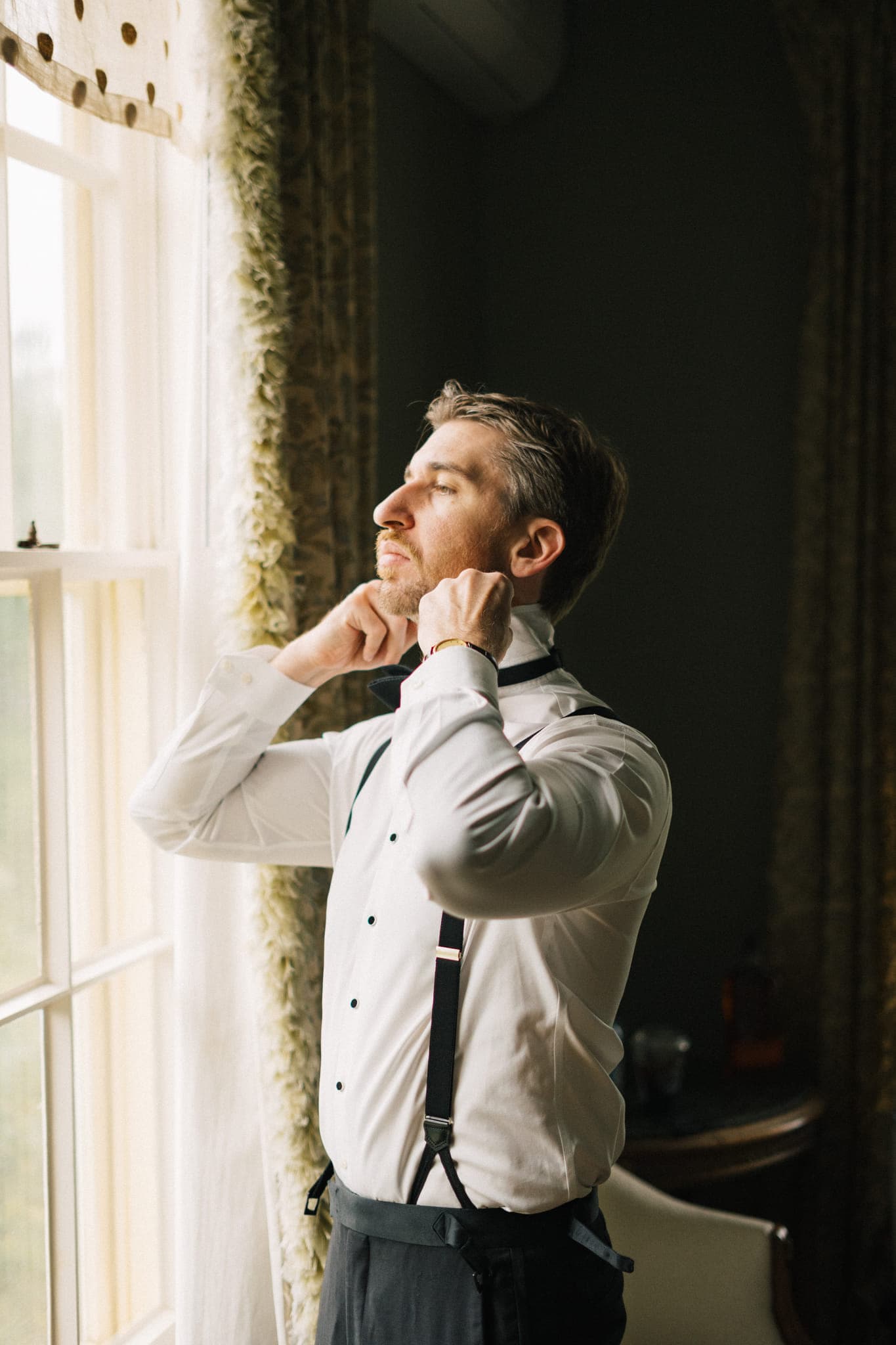 A man in a white shirt and suspenders adjusts his bow tie near a window with curtains.