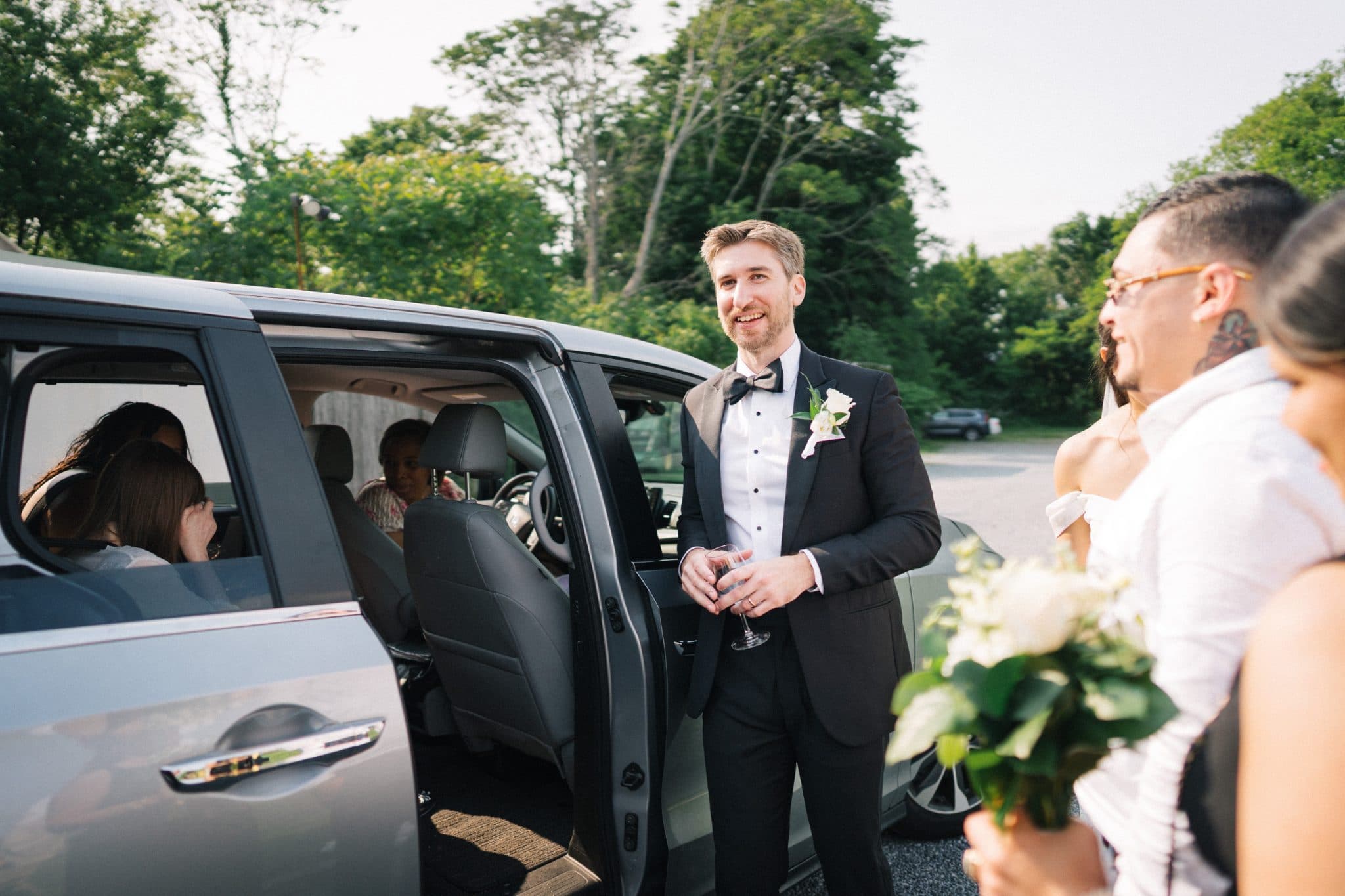 A man in a suit stands beside an open car door, holding a glass, while others gather nearby.