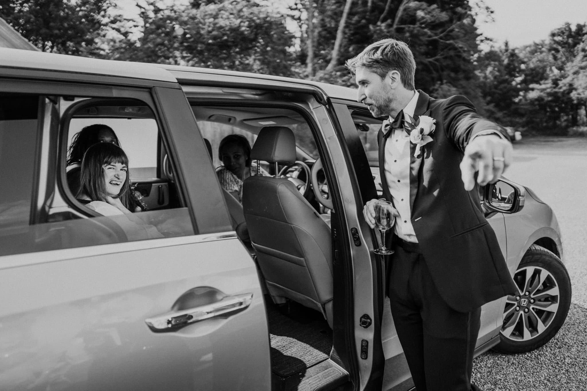 A man in a suit exits a car, holding a drink, while another person sits inside the vehicle.