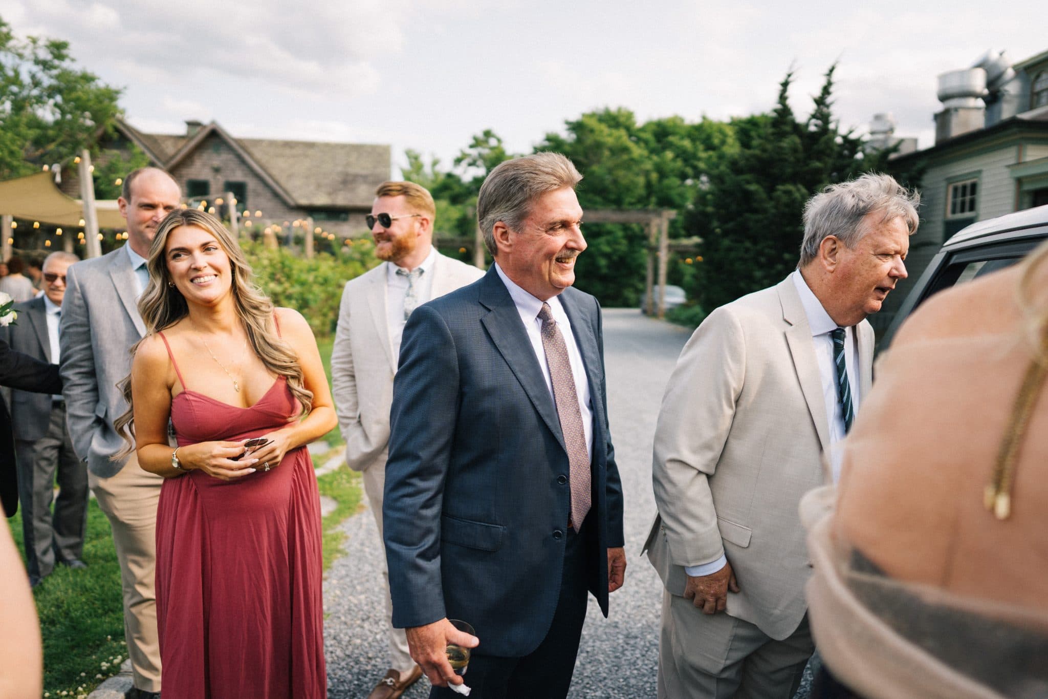 A group of people in formal attire stands outdoors, smiling and conversing near a building with a vineyard in the background.
