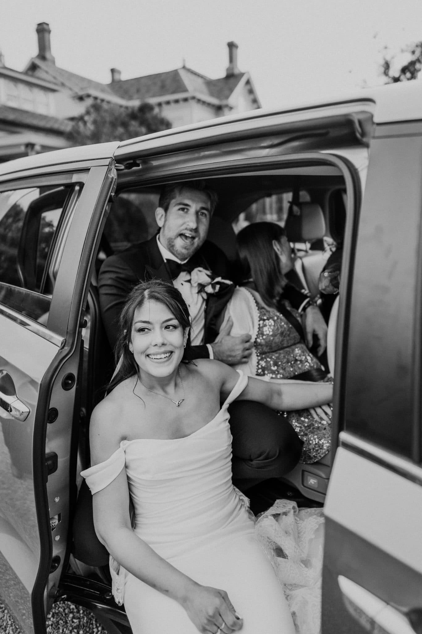 A bride in a white dress smiles as she sits in the back of a car with a man in a suit and another woman in a patterned dress.