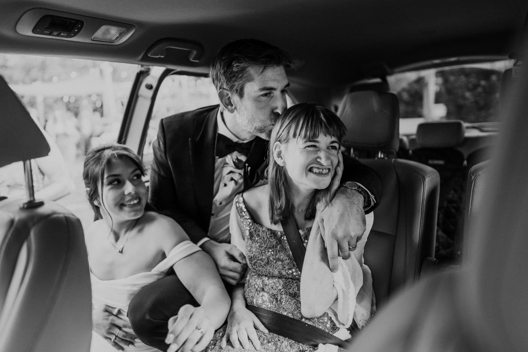 Three people in a car, one in a suit and bow tie, another in a dress, and the third in a white top, with the suited person leaning towards the third person.