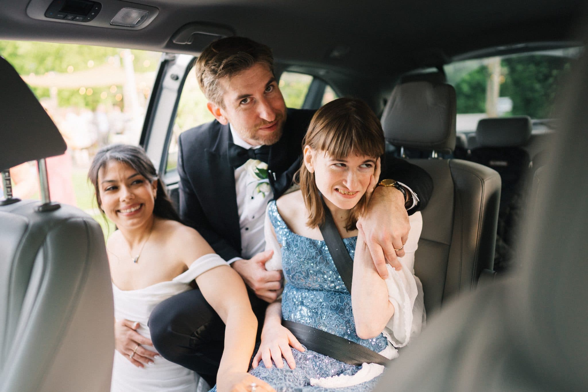 Three people in formal attire sit in a car, smiling and embracing.