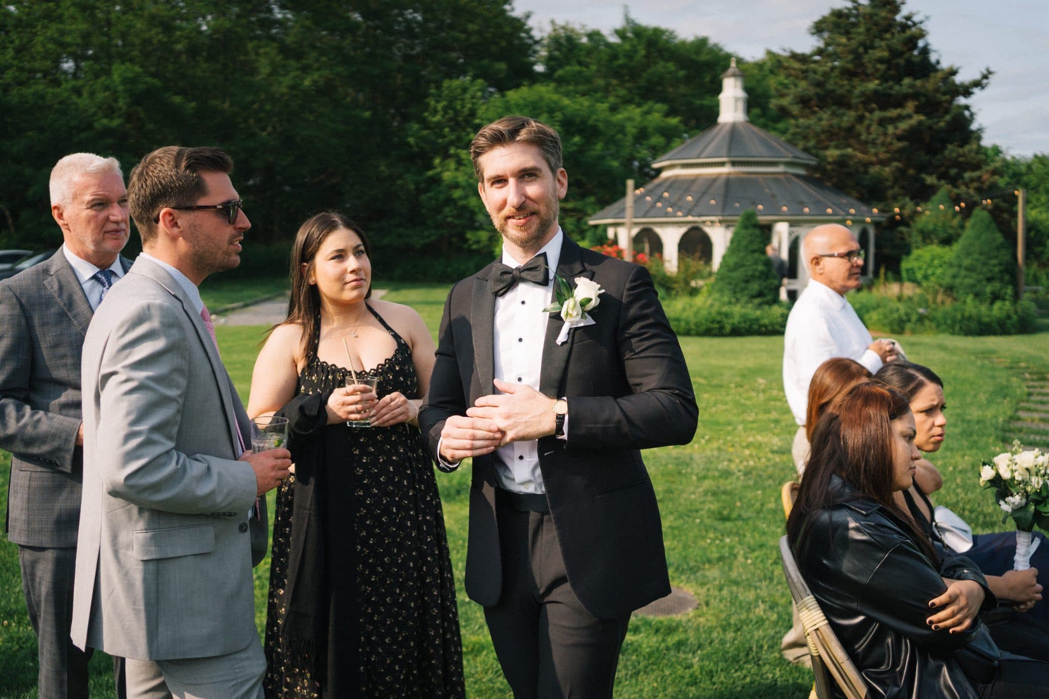A man in a tuxedo stands in a grassy area with guests, some holding drinks, near a gazebo.