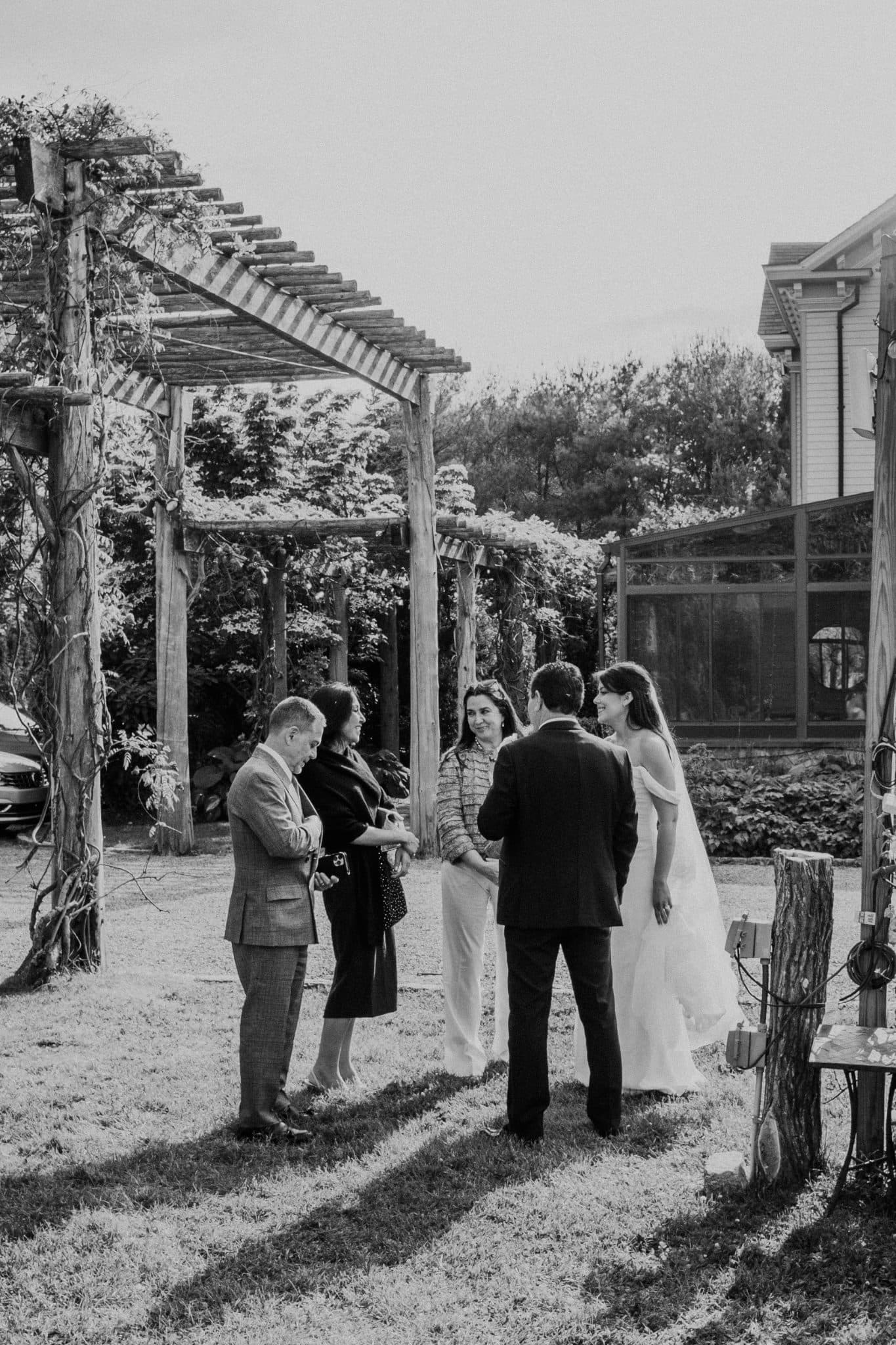 A black-and-white wedding scene with a bride in a white dress, groom in a suit, and guests in formal attire standing under a wooden pergola.