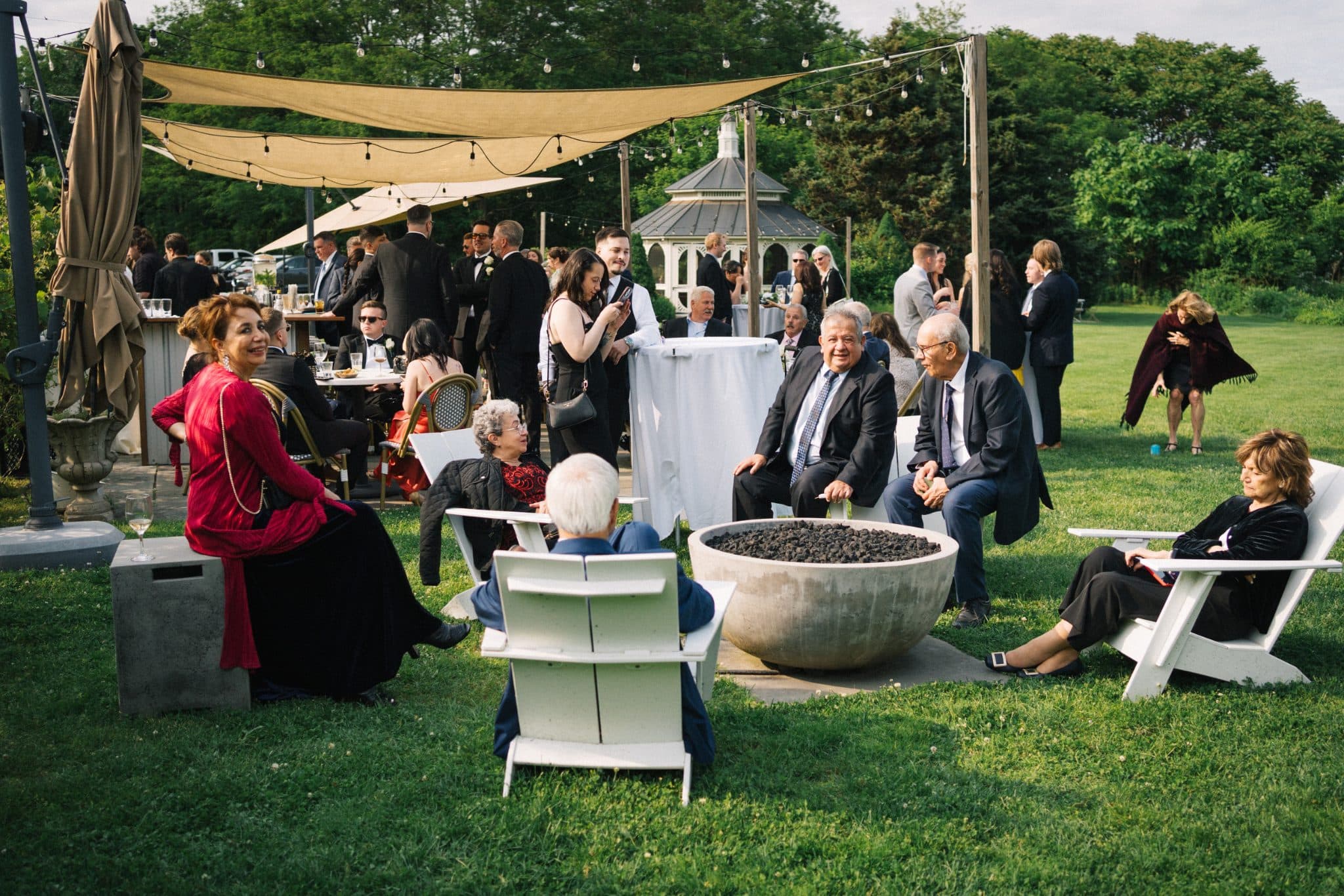 A group of people in formal attire gather outdoors under a canopy, sitting and standing around a fire pit and tables.