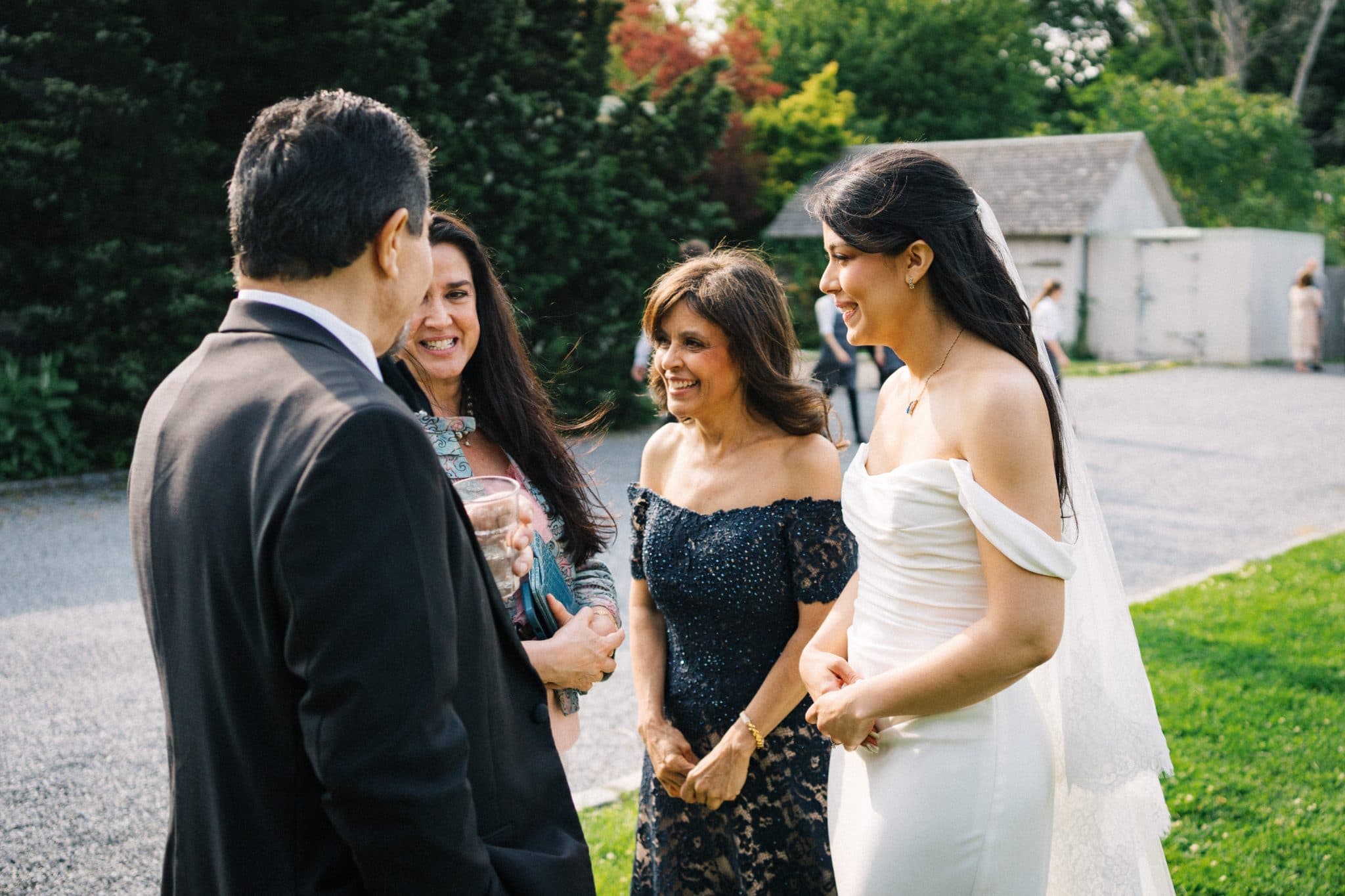 A bride in a white dress stands with a man in a suit and two women in elegant dresses, smiling and talking in a garden setting.