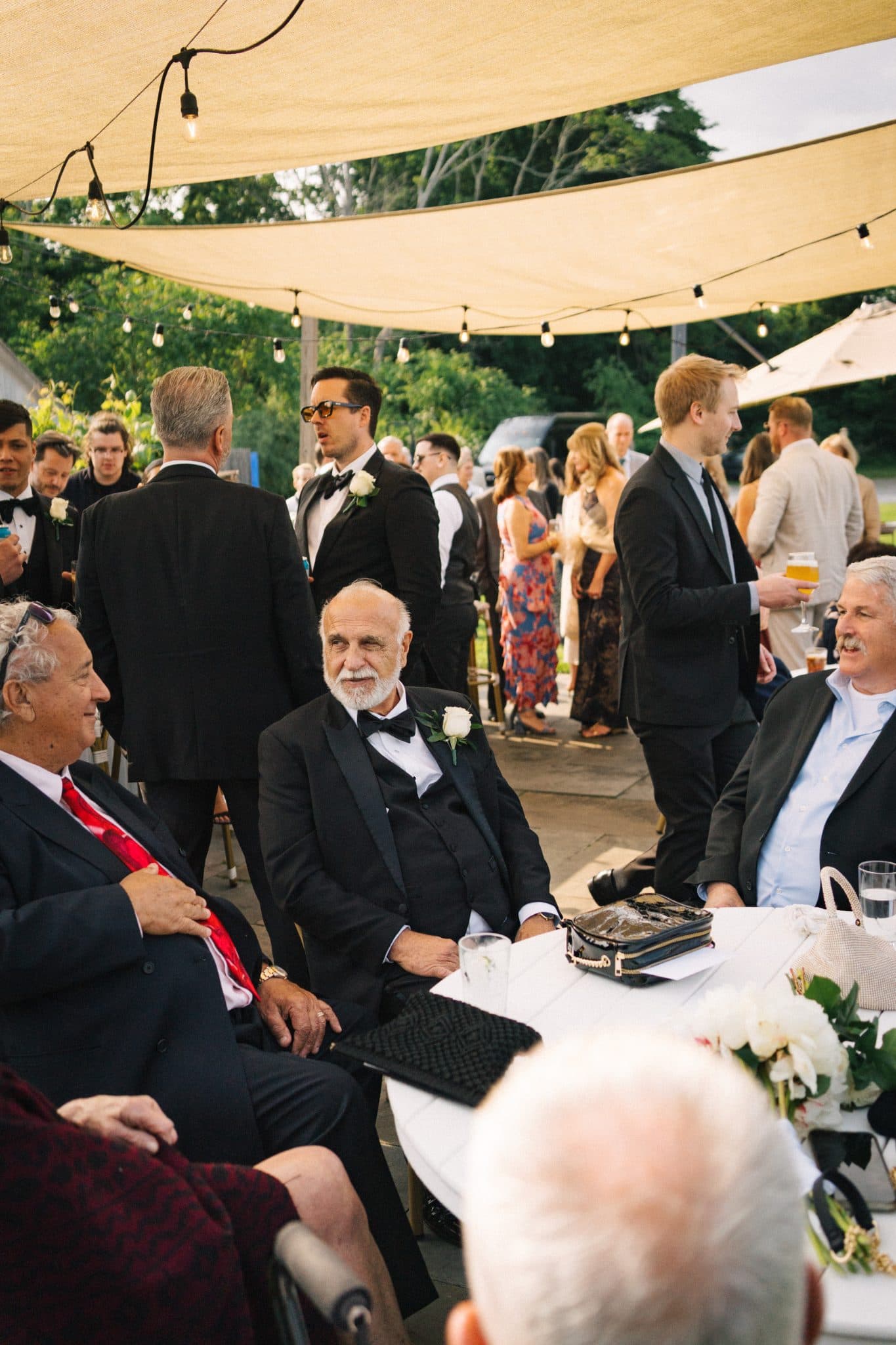 A group of people dressed formally, some in suits, are gathered under a canopy at an outdoor event.