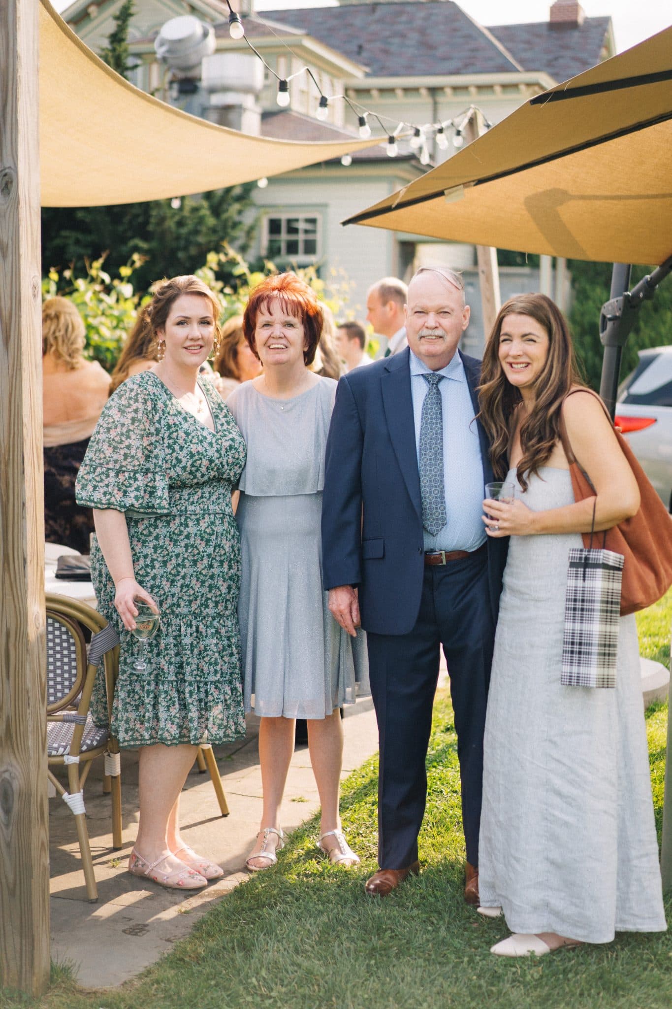 Four people stand together outdoors, smiling, with a house and string lights in the background.