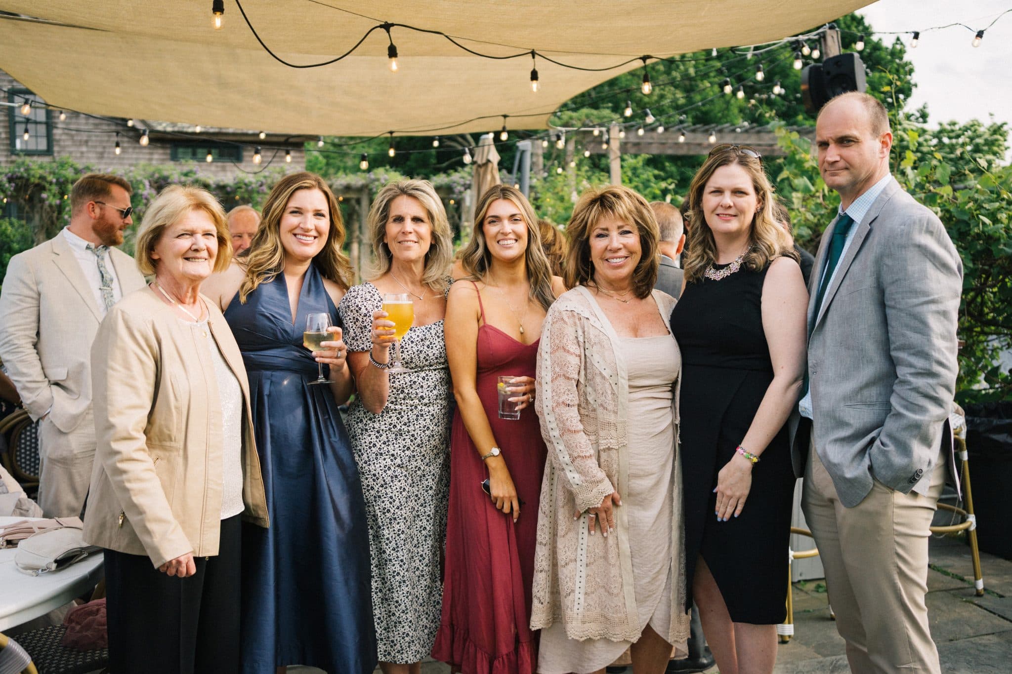 A group of people, dressed in formal attire, stands together under a canopy with string lights, smiling and holding drinks.