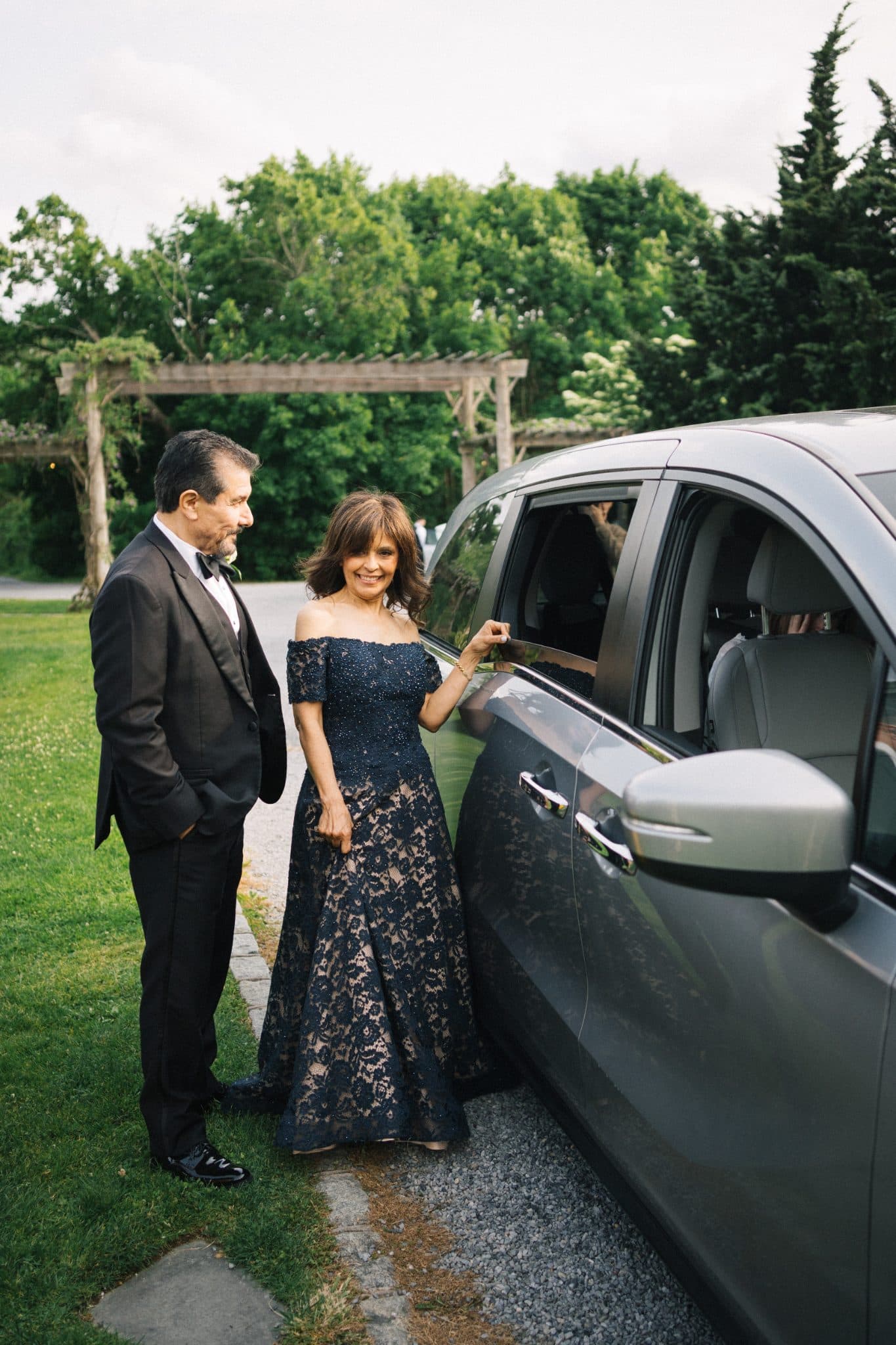 A woman in a lace gown and a man in a suit stand beside a car on a grassy path.