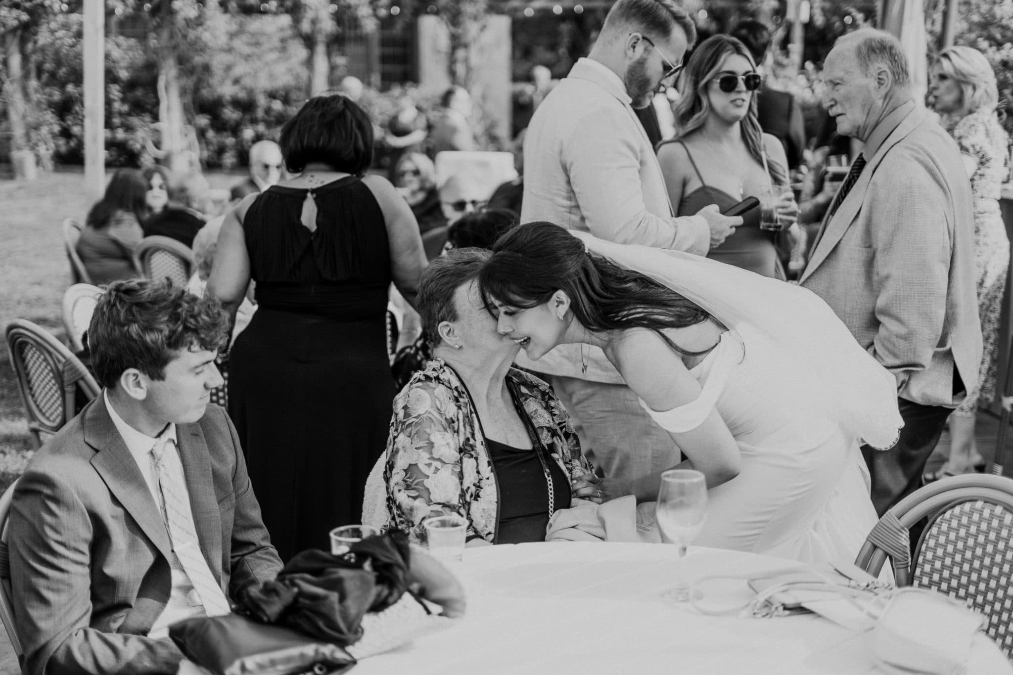 A bride leans over a table to speak to a seated woman at a wedding reception.