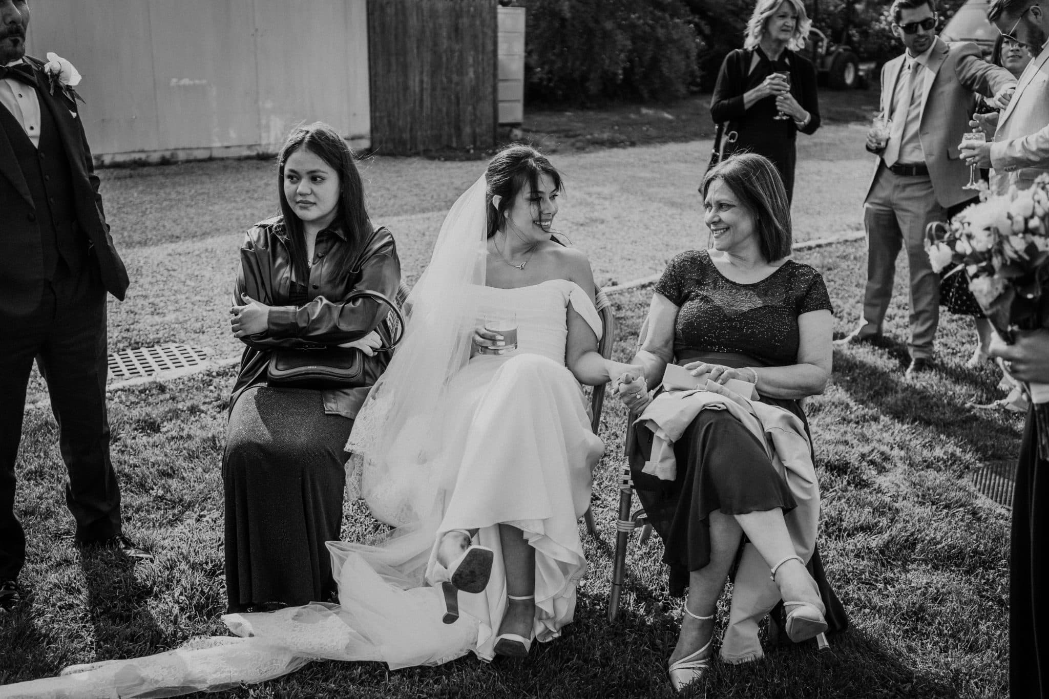 A bride in a white wedding dress sits between two women on a grassy lawn, surrounded by other guests.