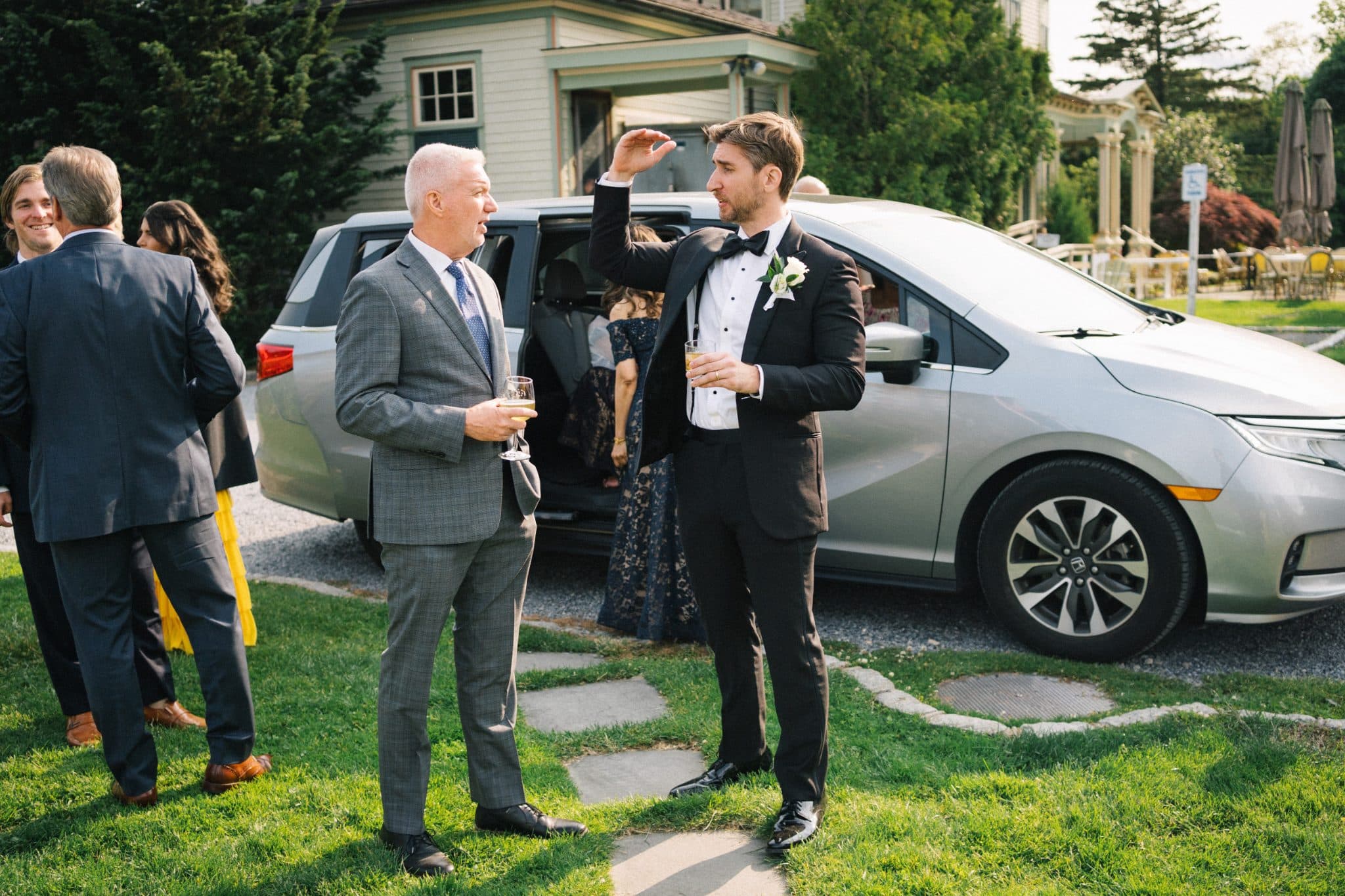A man in a tuxedo holds a drink, talking to another man in a suit near a silver van and a house with greenery.