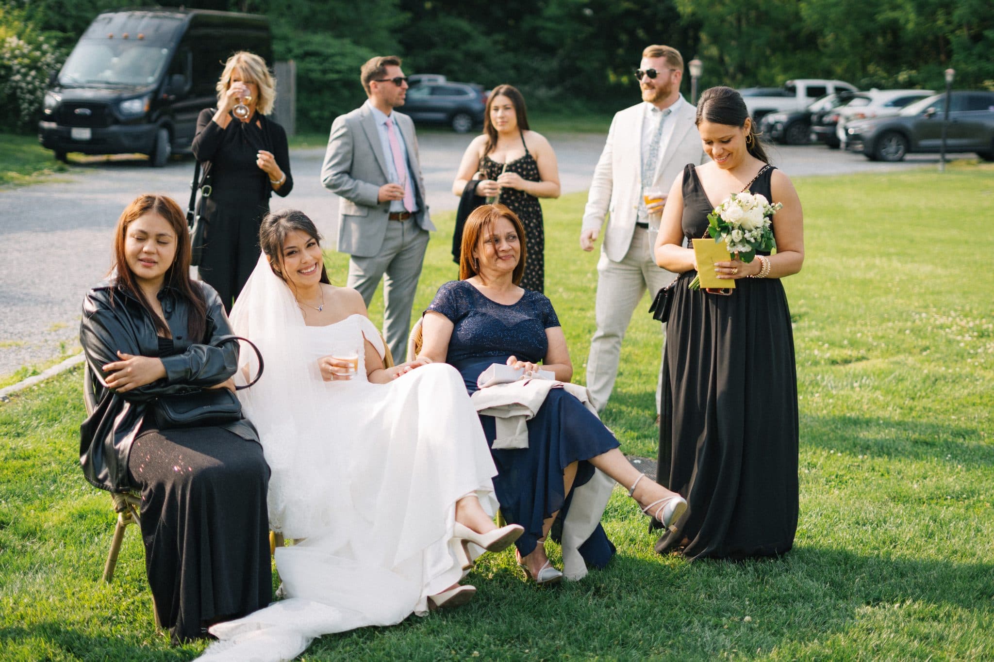 A bride in white sits on a chair with guests in formal attire, some holding drinks, on a grassy lawn.