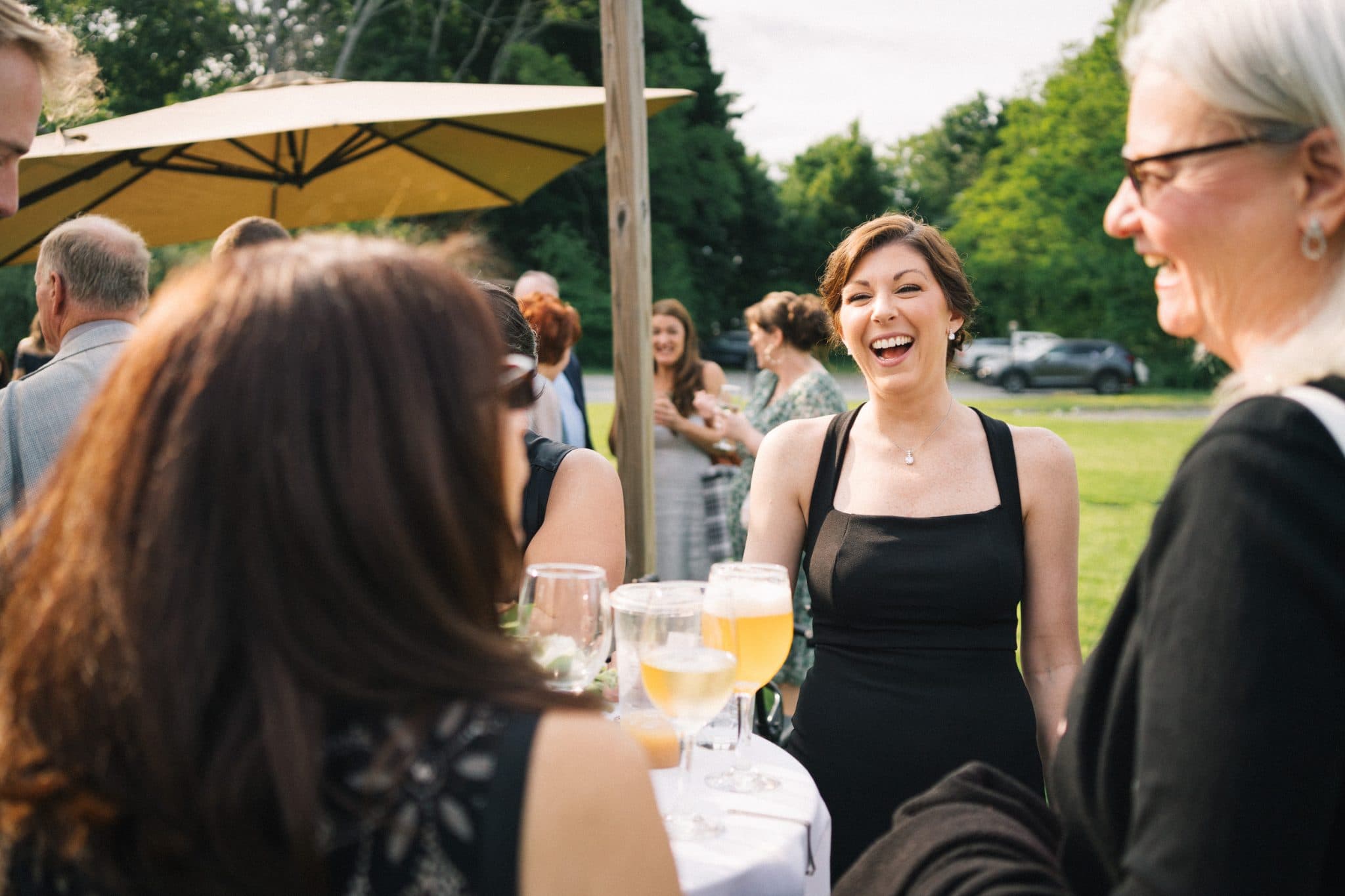 A woman in a black dress laughs while standing near a table with drinks and a yellow umbrella in the background.