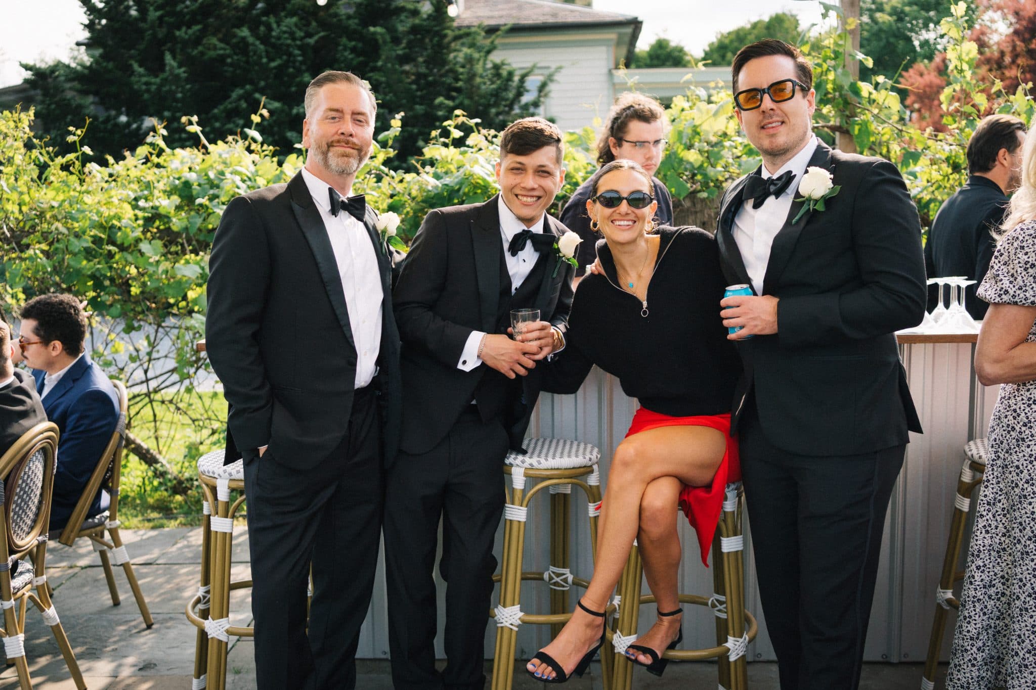 Four people in formal attire pose together outdoors with a backdrop of greenery and a building.