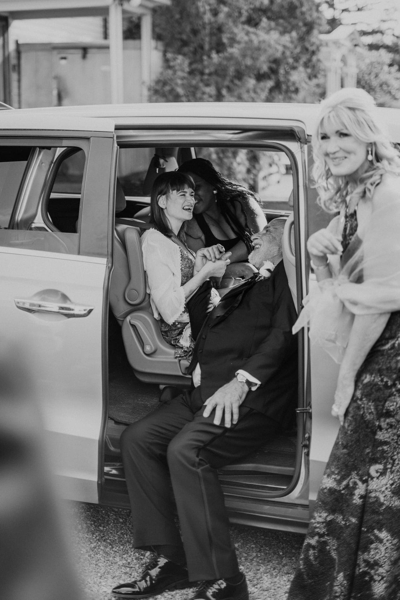 A man in a suit sits in the back of a van, holding hands with a woman in a white top, while another woman in a floral dress stands nearby.
