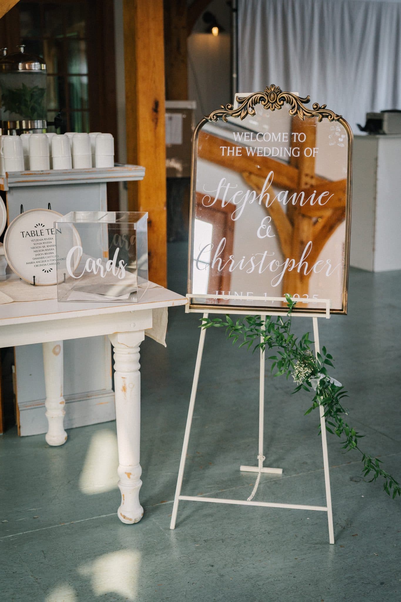 A wedding welcome sign on a stand with greenery, next to a table with a sign for cards and cups.