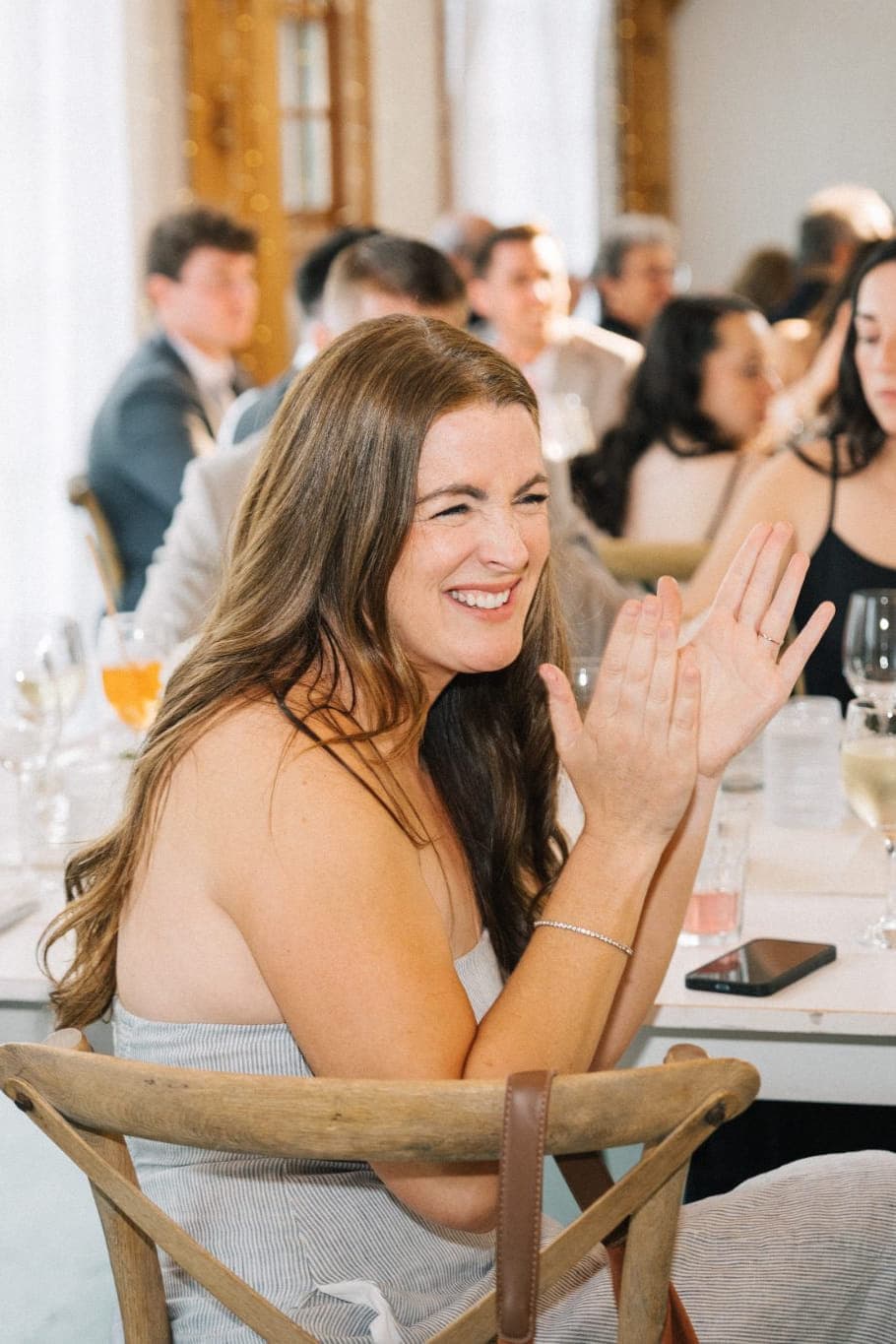 A woman in a white dress claps her hands at a table with other guests, some clapping and smiling, at a formal event.