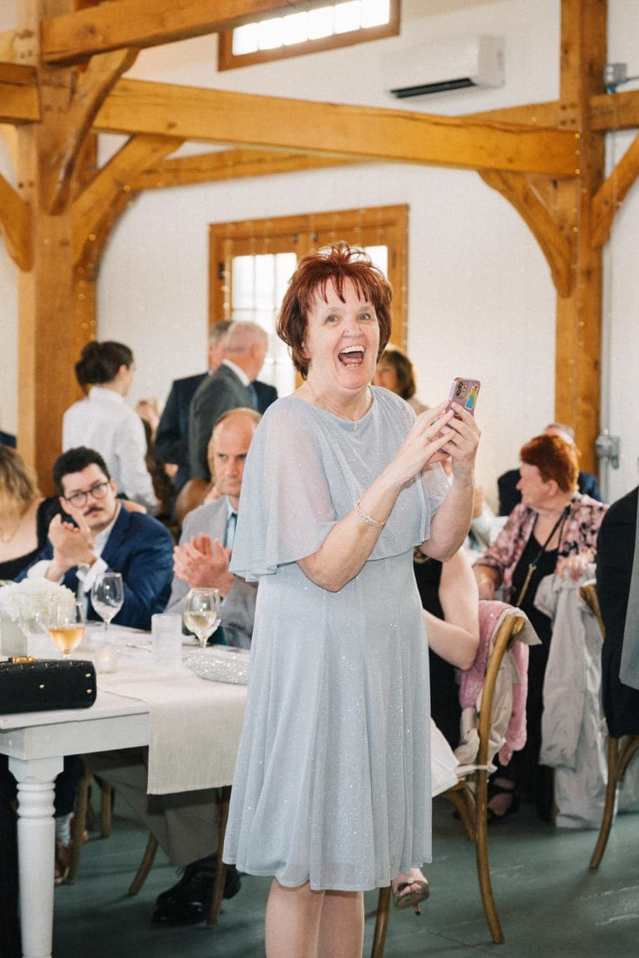 A woman in a light blue dress stands smiling, holding a phone, surrounded by seated guests at a wedding reception.
