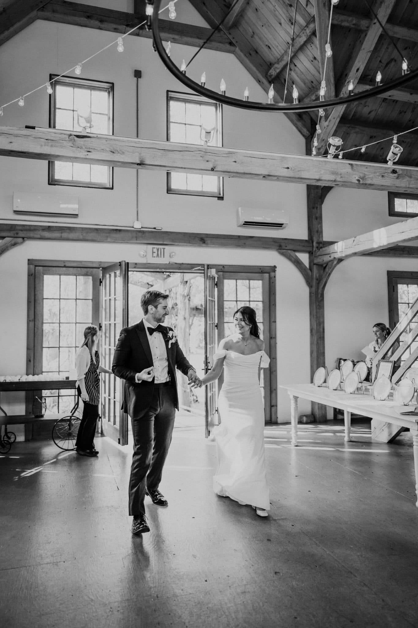 A bride and groom walk hand in hand through a barn, smiling and holding hands.