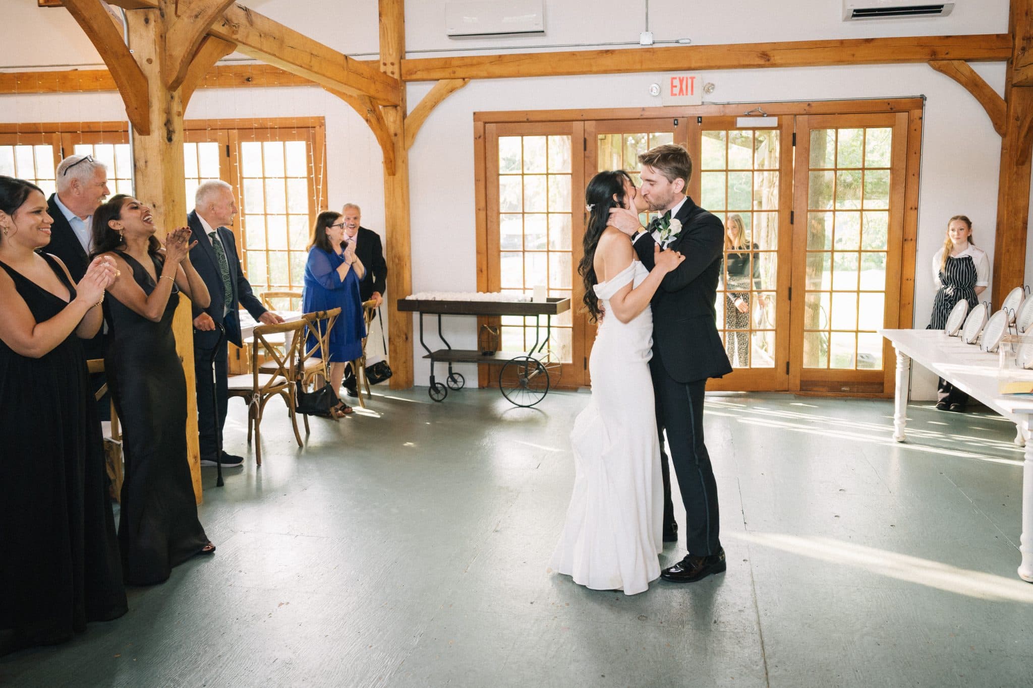A bride and groom share a kiss as guests clap and watch in a wooden-beamed room.