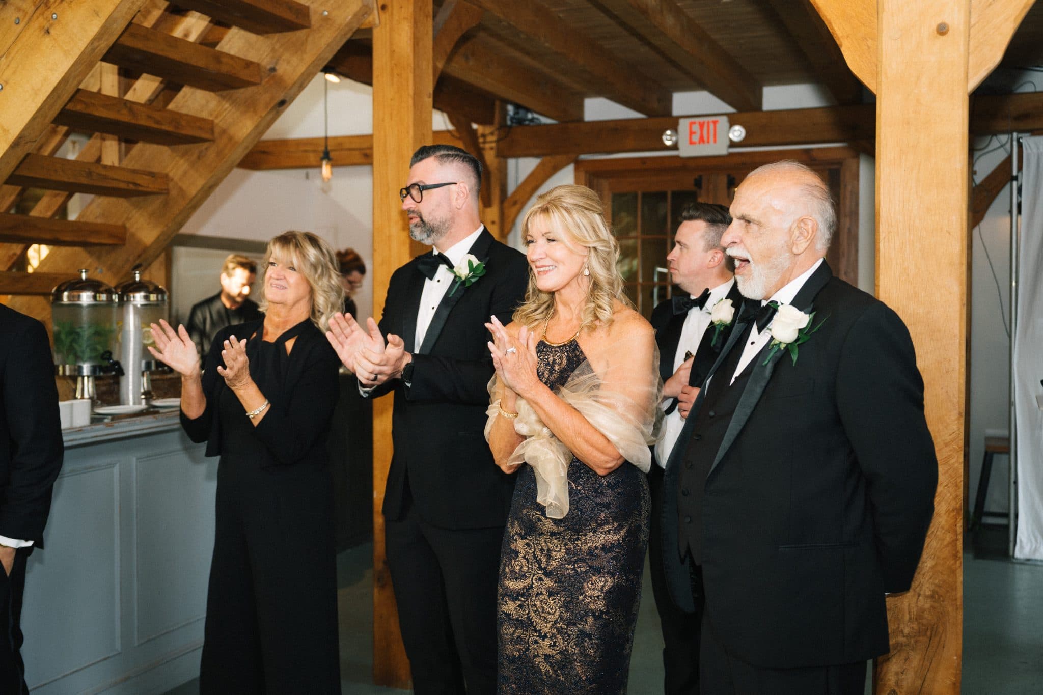 A group of people in formal attire claps and smiles in a wooden-beamed room.
