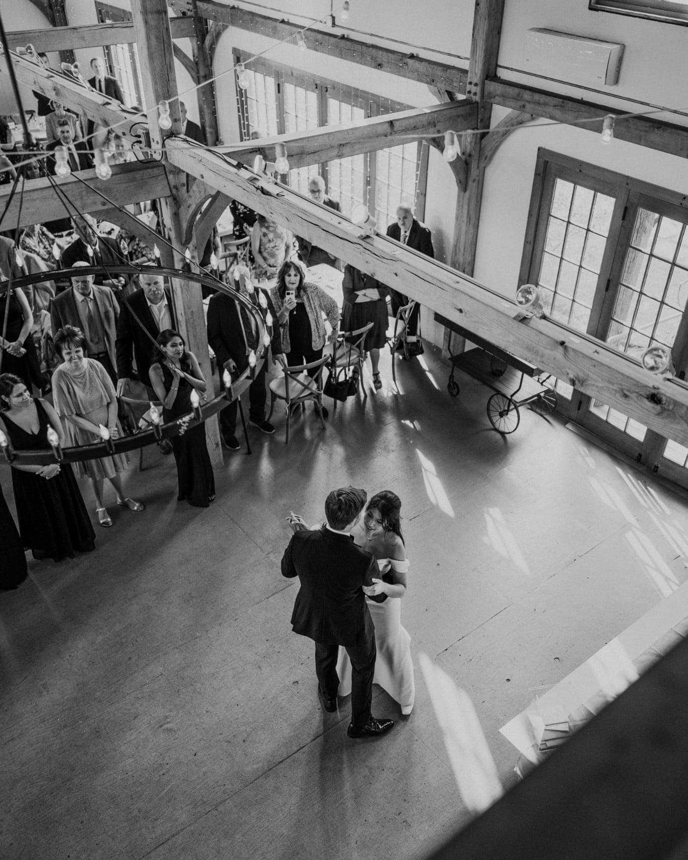 A bride and groom share a kiss on a dance floor surrounded by guests in a large, rustic venue with wooden beams and windows.