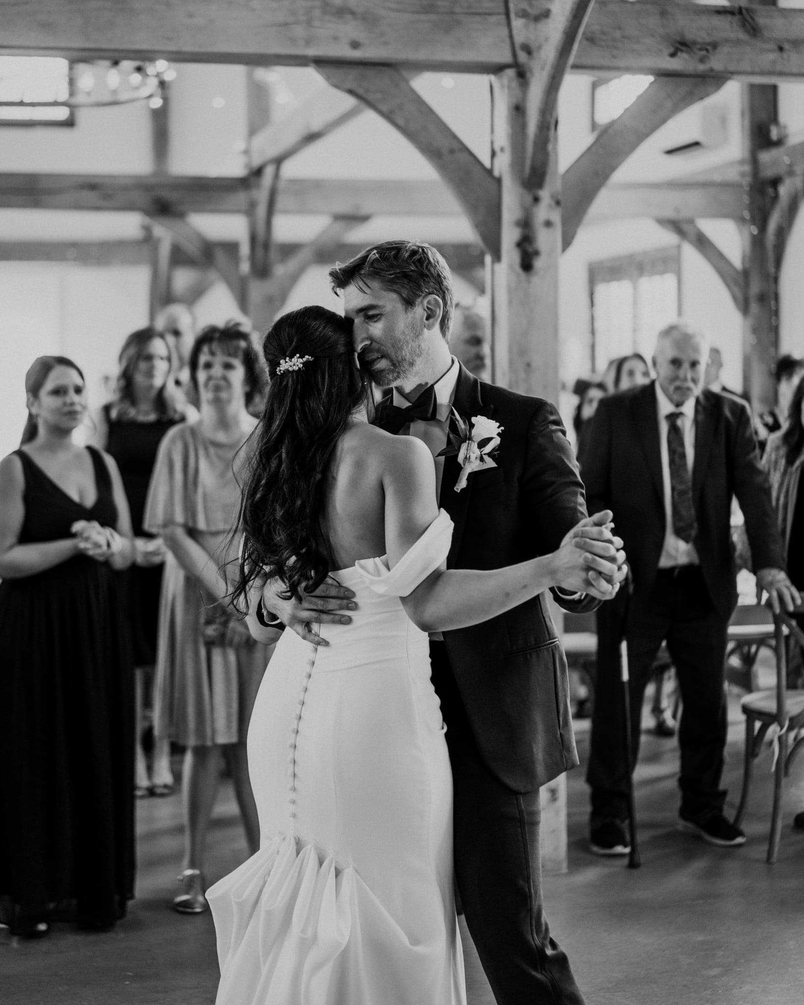 A bride and groom share a dance in a rustic barn setting, surrounded by guests.