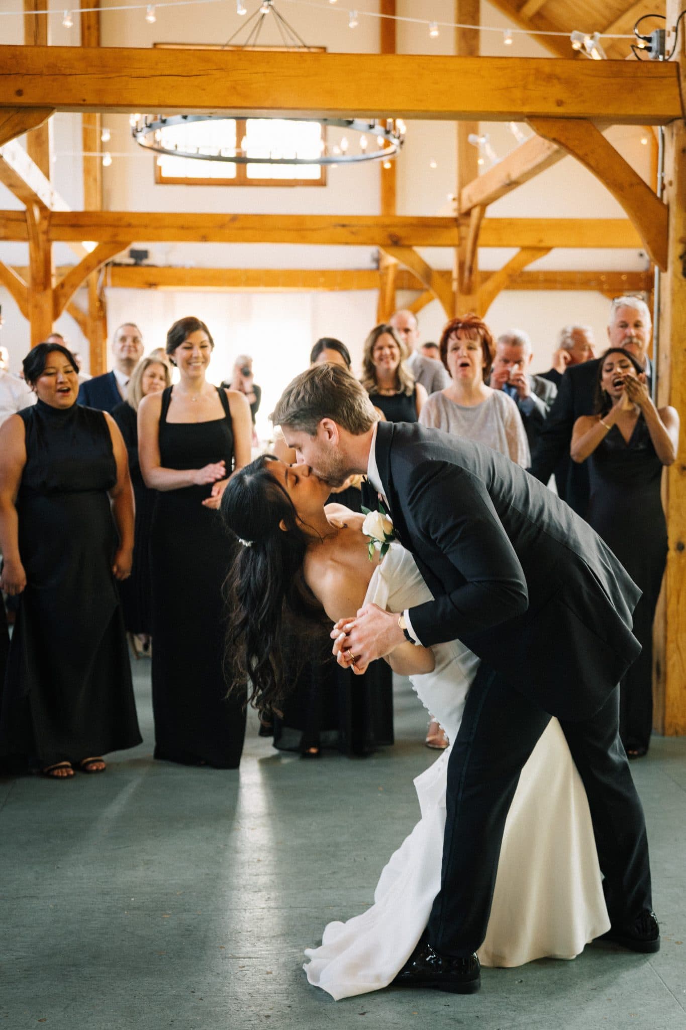 A bride and groom share a kiss in a barn-style venue surrounded by guests.