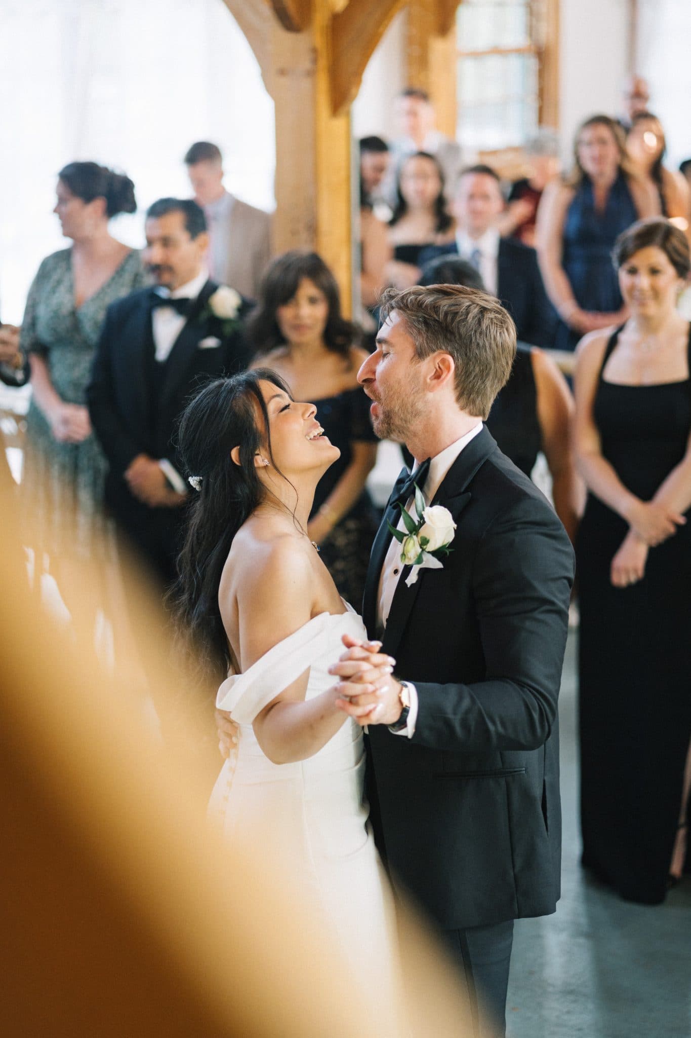 A bride in a white dress and a groom in a black suit hold hands, smiling and looking at each other, with guests in formal attire in the background.